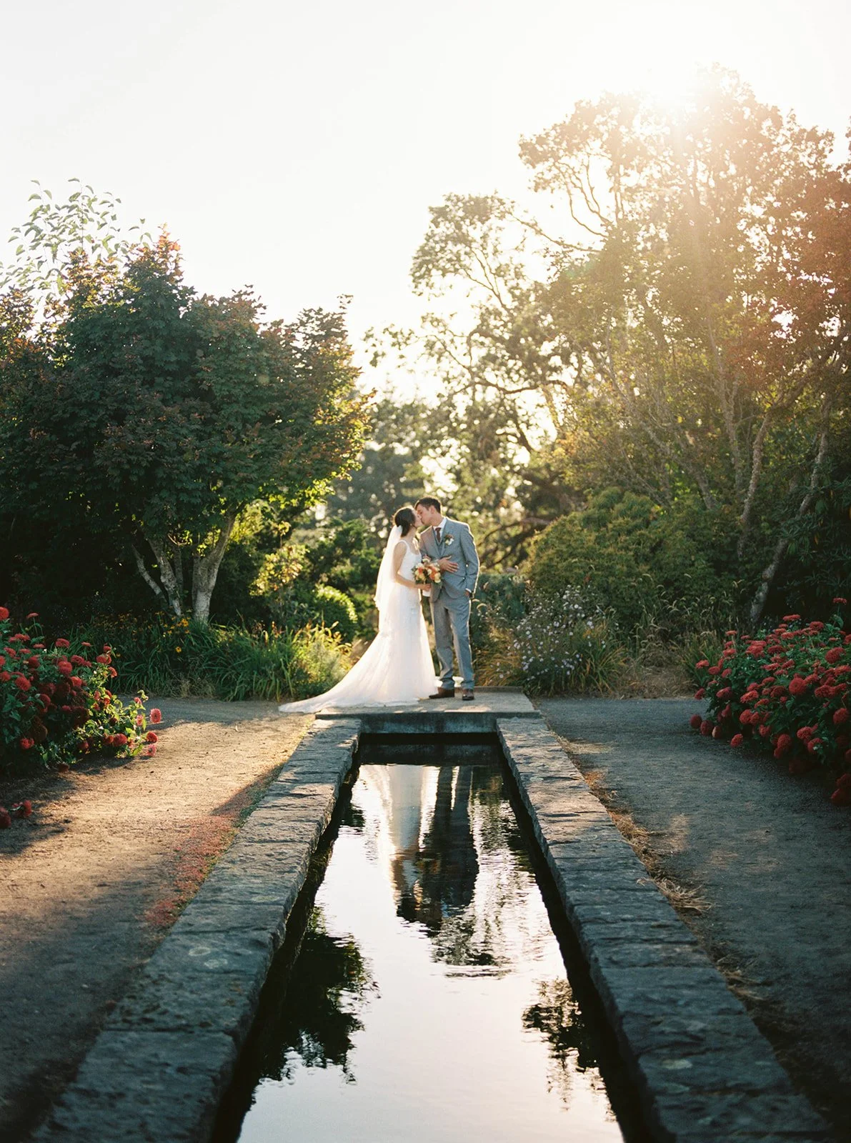 Bride and groom kissing beside a reflecting pool surrounded by gardens in warm evening light.