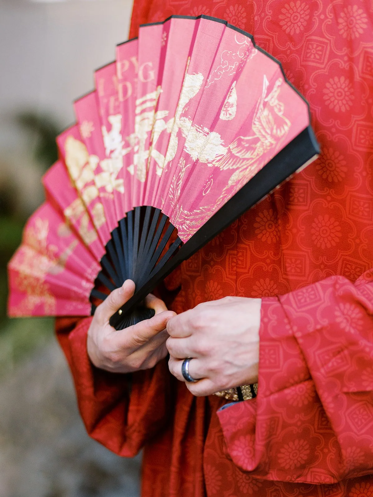 lose-up of the groom's hands holding a red and gold "Happy Wedding" Chinese fan against his red patterned ceremonial robe, wedding band visible