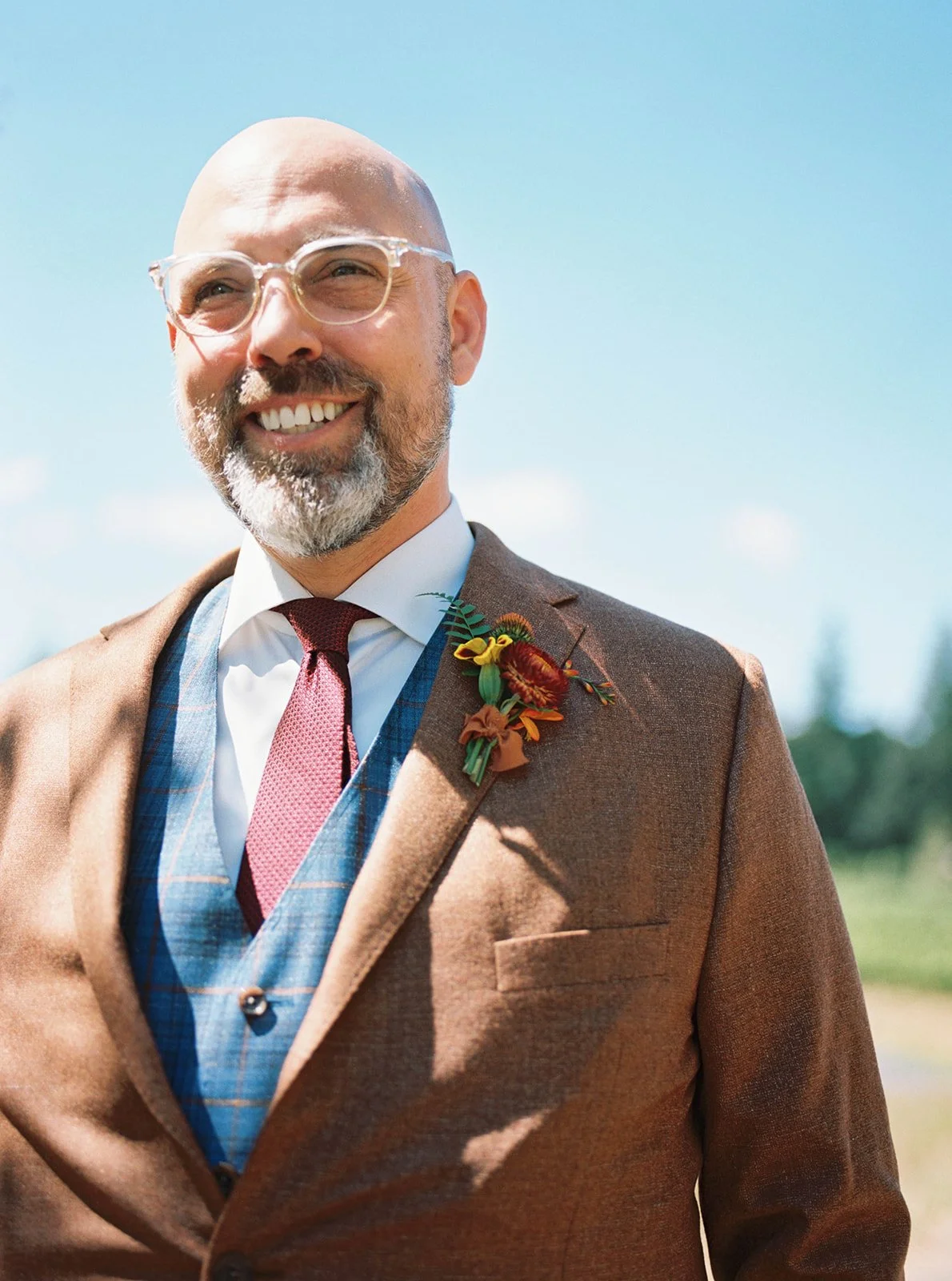Close-up of groom in brown suit with boutonniere, smiling outdoors in natural light