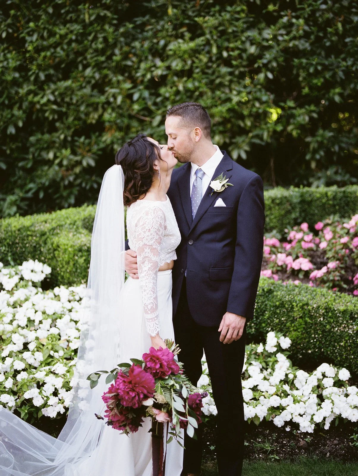 Bride and groom share a quiet kiss during their Lewis and Clark Estate Gardens wedding, surrounded by manicured hedges and bold magenta florals.