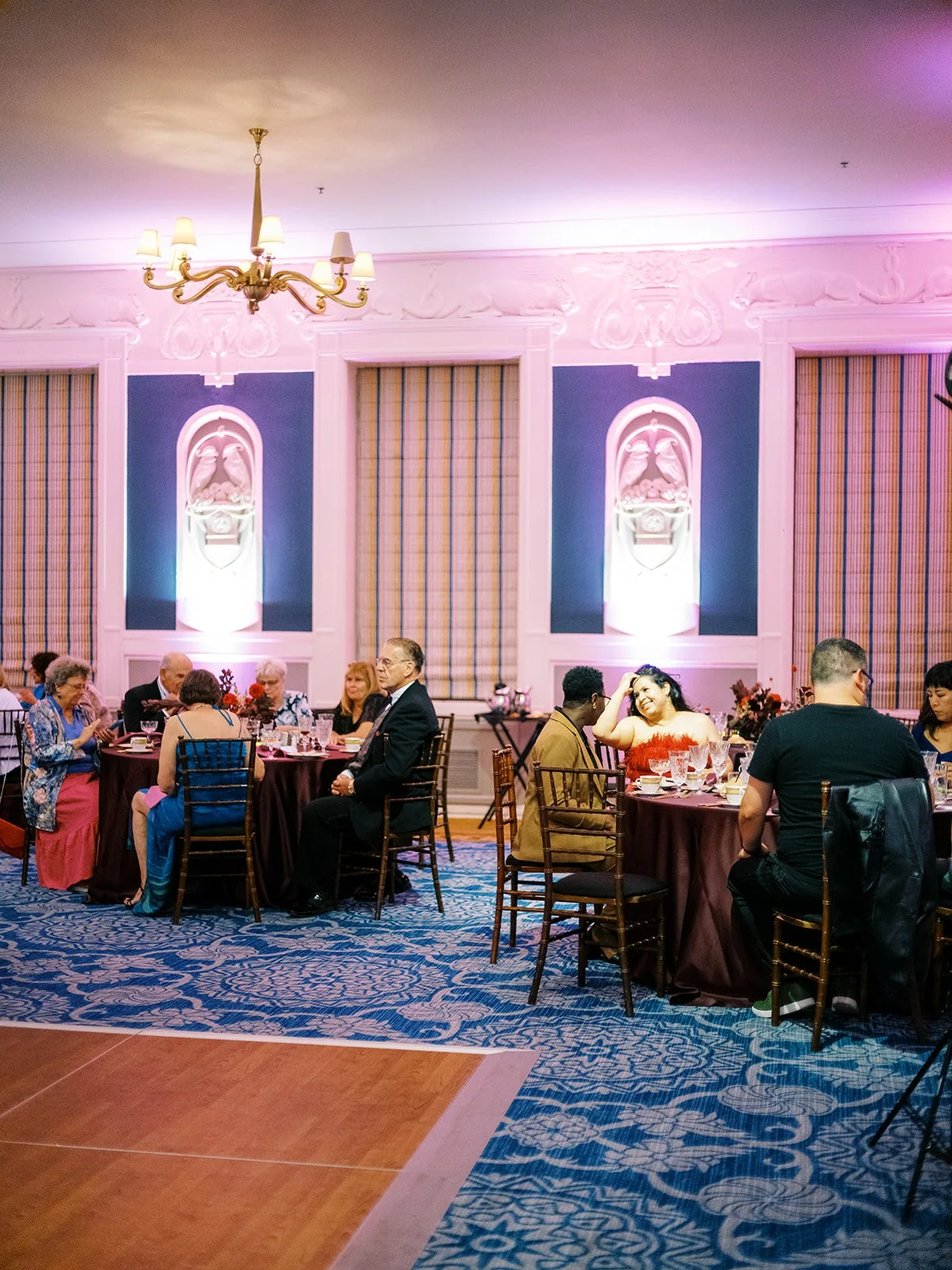 Wedding guests seated at round tables in a decorated reception hall with chandeliers and pink uplighting.