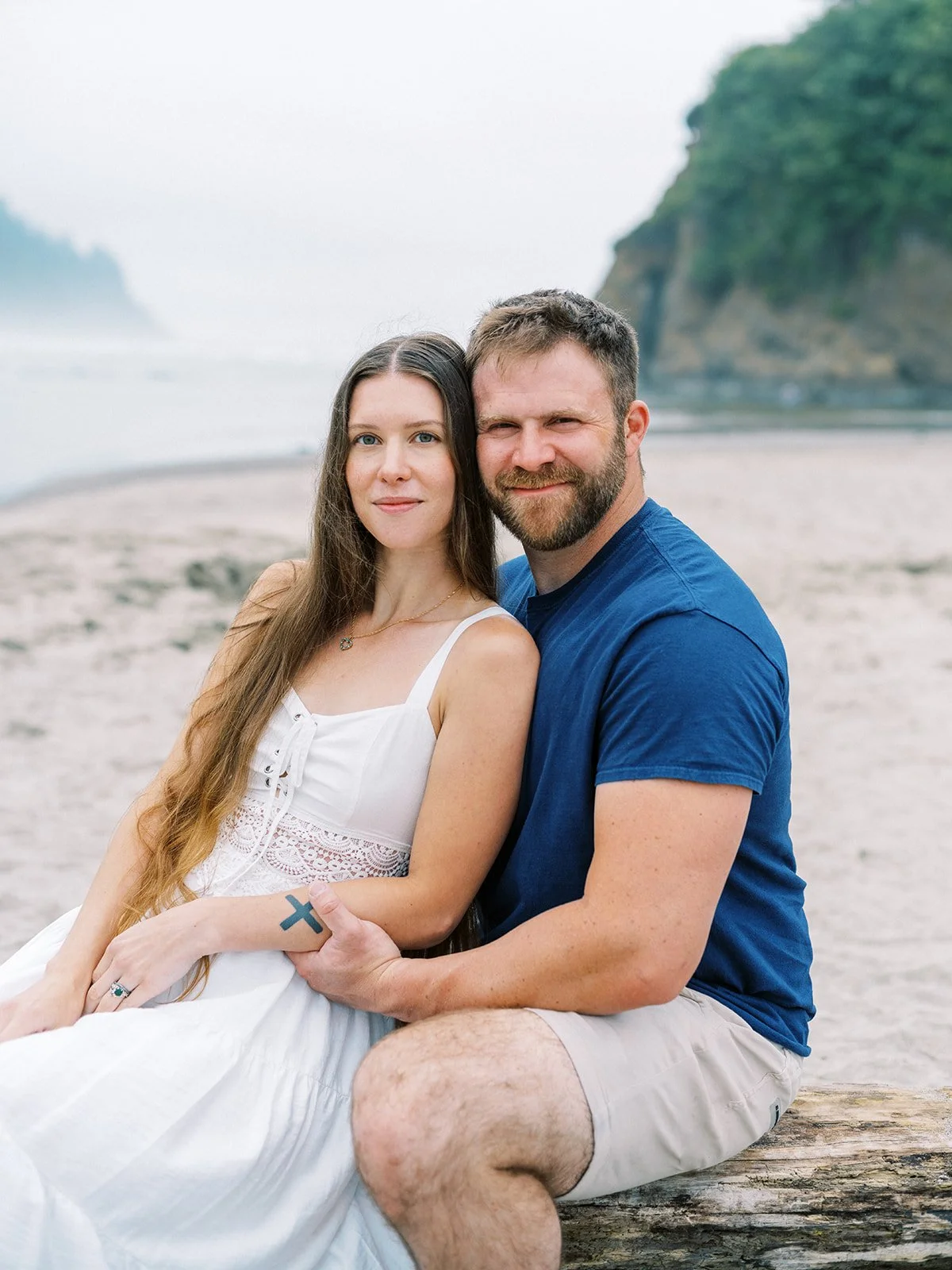 Couple sitting on driftwood log on quiet beach with soft fog and rocky coastline behind them.