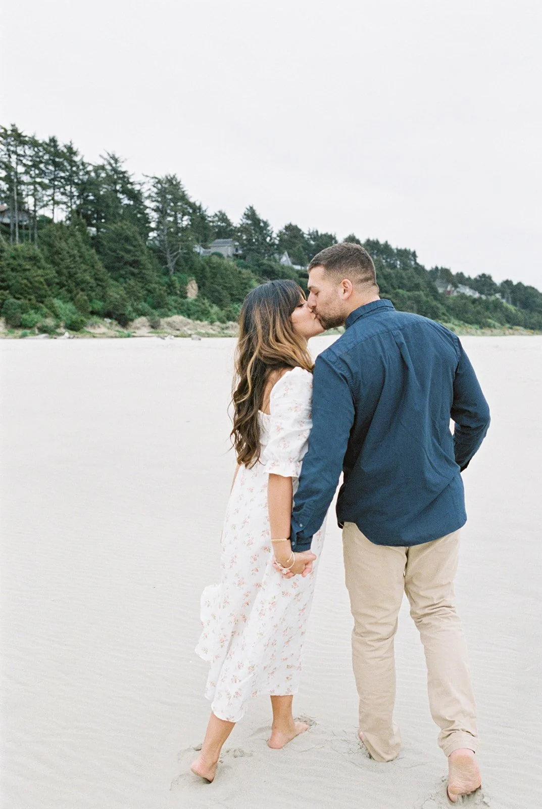 Couple holding hands and kissing on beach with coastal trees in background
