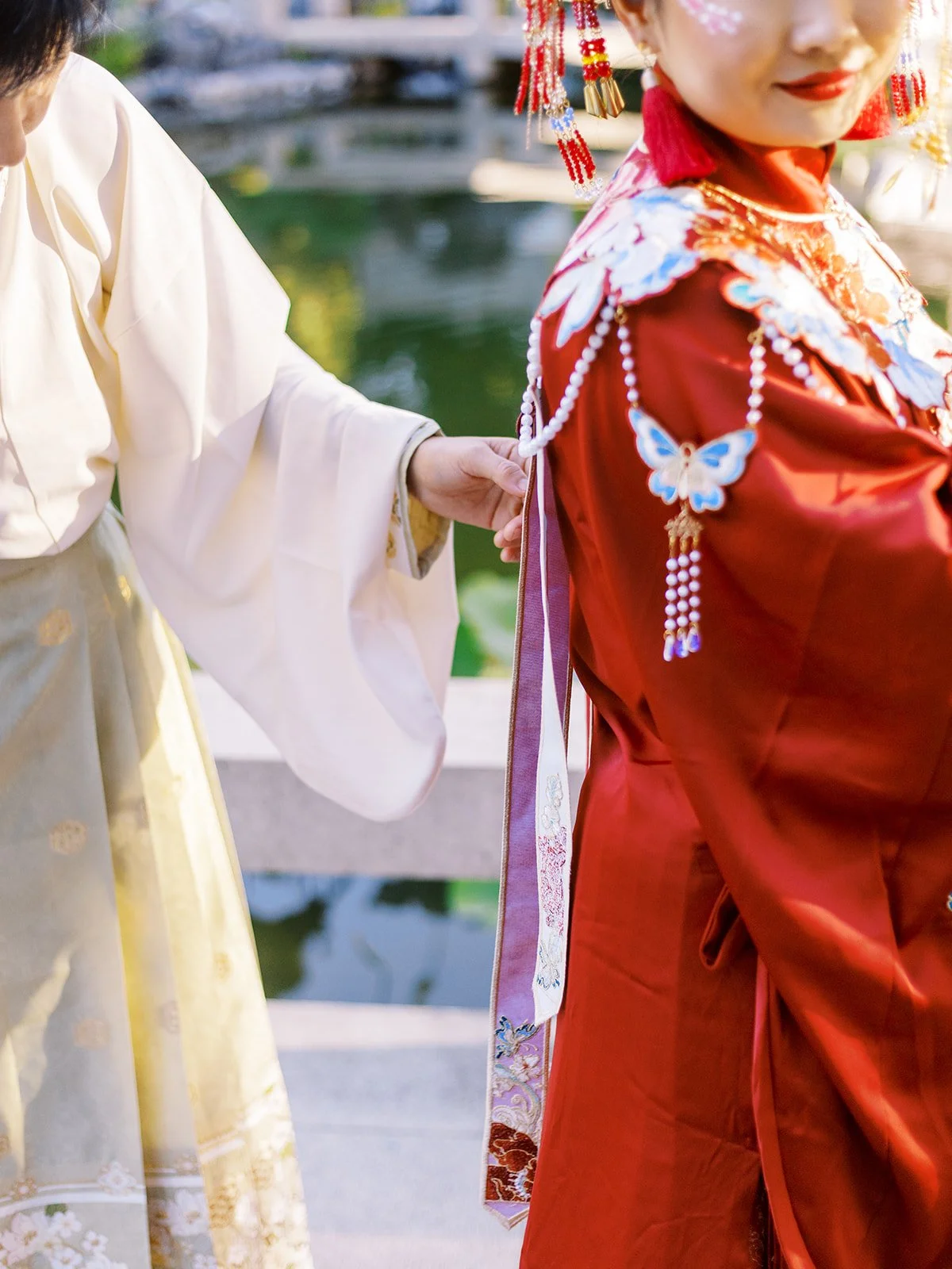 Person tying the back of a red traditional wedding robe with embroidered details beside a garden pond