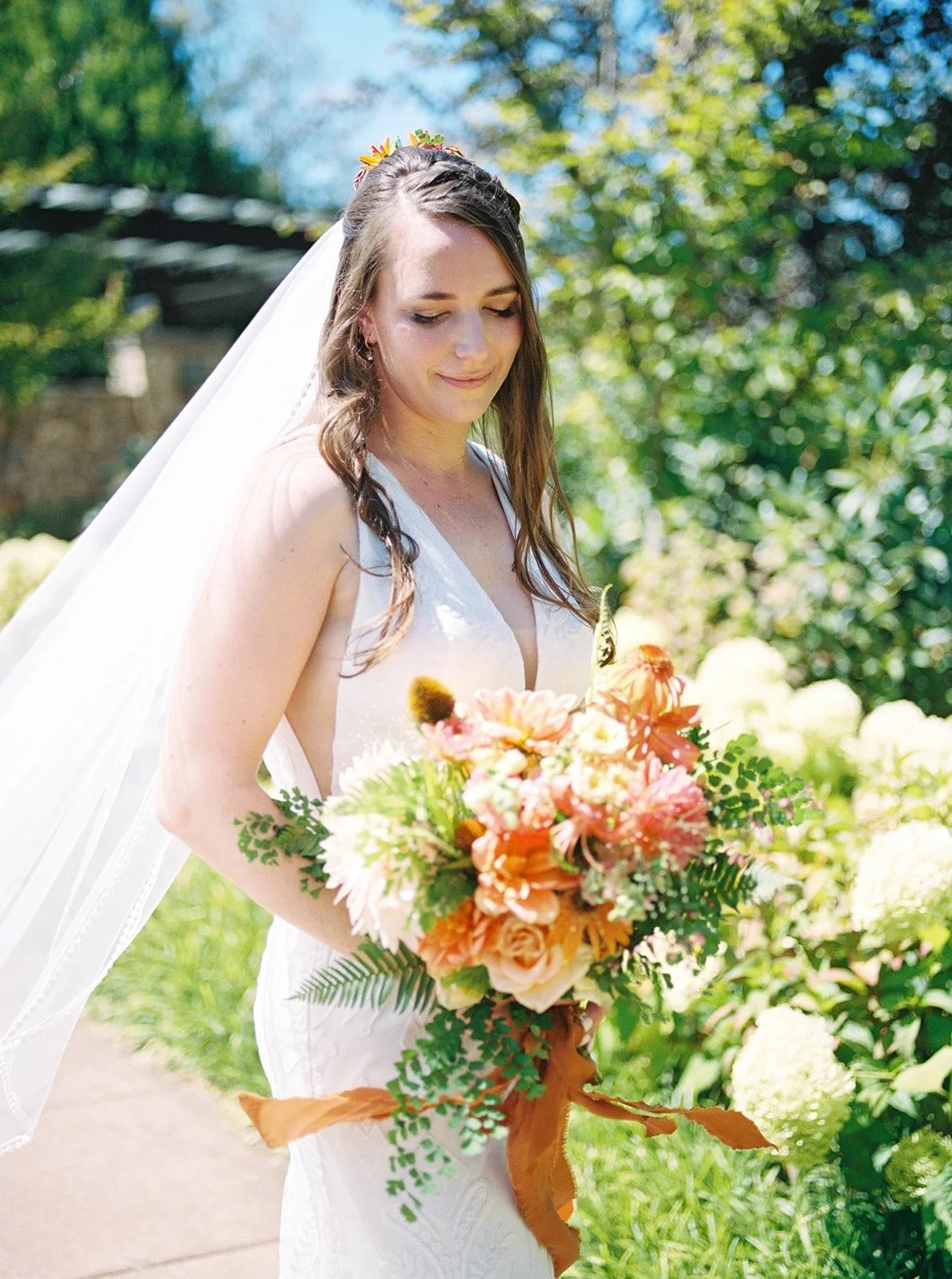 Bride with veil looking down at bouquet of orange, peach, and cream flowers in garden