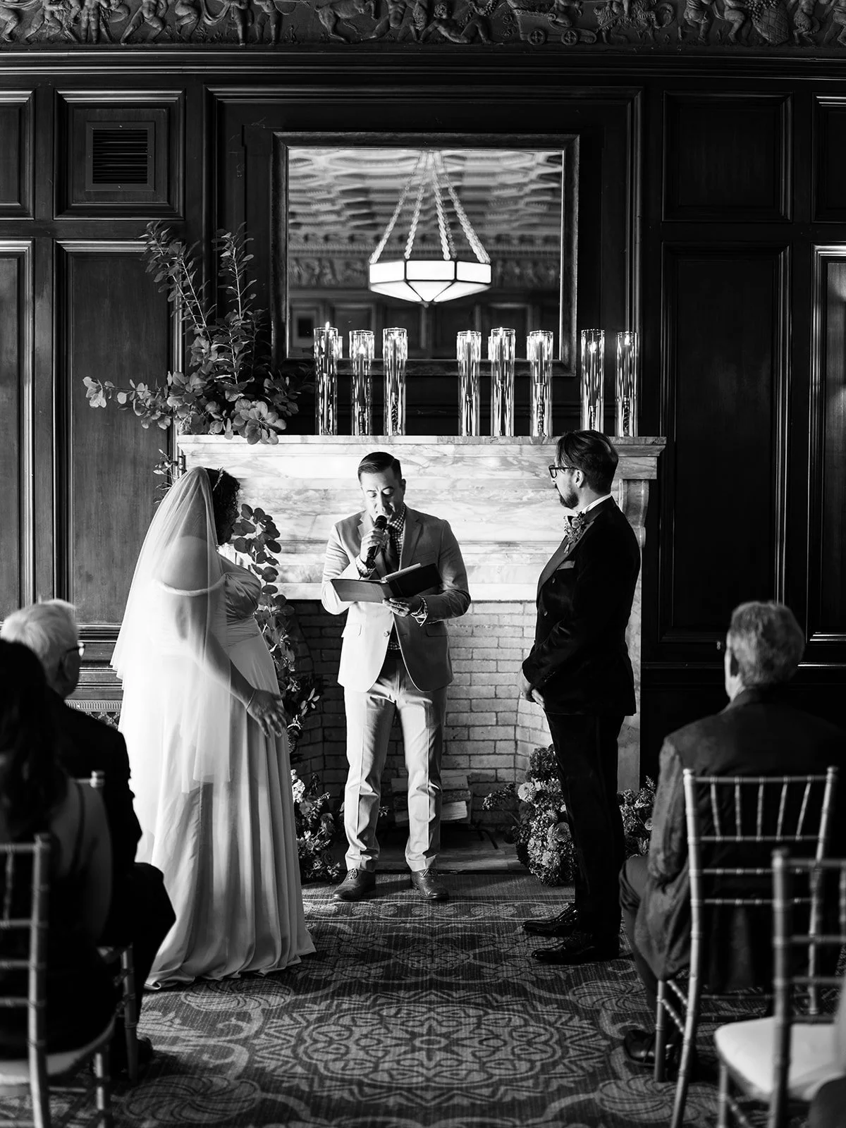 Black and white image of couple holding hands during wedding ceremony at fireplace altar.