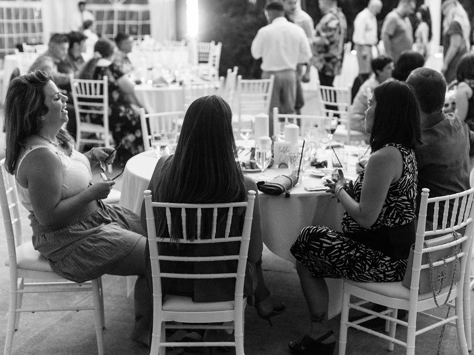 Wedding guests seated and socializing at round reception tables under a tent in black-and-white.