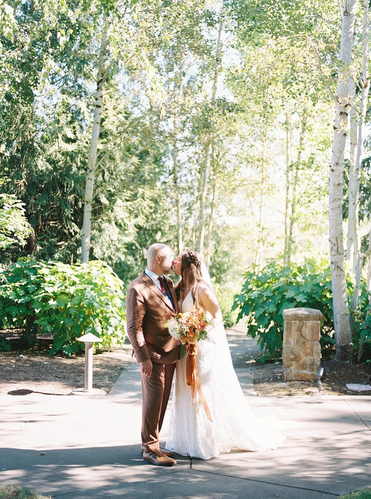 Bride and groom kiss beneath tall trees in a garden setting, soft sunlight filtering through the leaves.