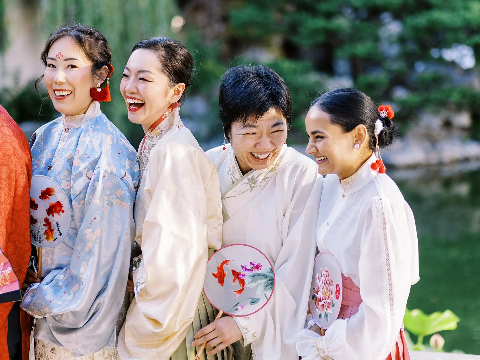 Four attendants in hanfu laughing together at Lan Su Chinese Garden, holding embroidered koi fish round fans against a lush garden backdrop