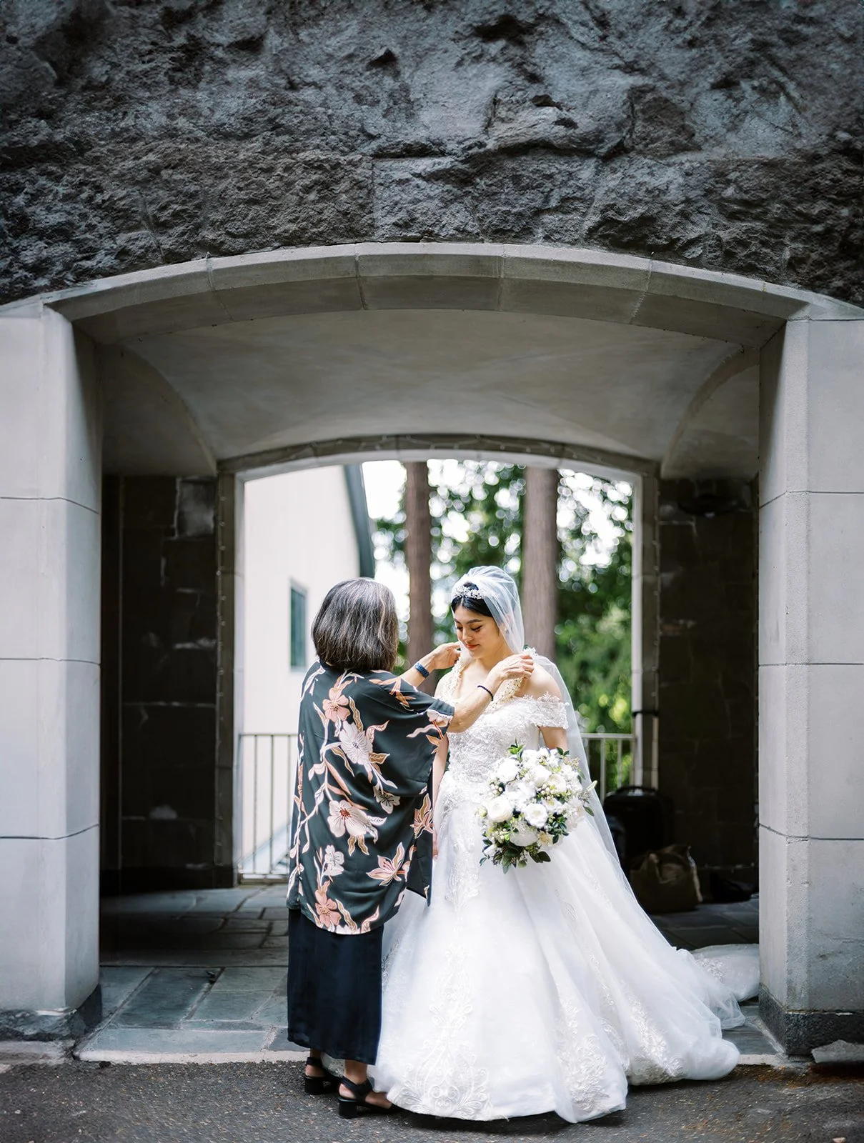 Woman adjusts a bride’s wedding dress while the bride holds a white bouquet beneath a stone archway.