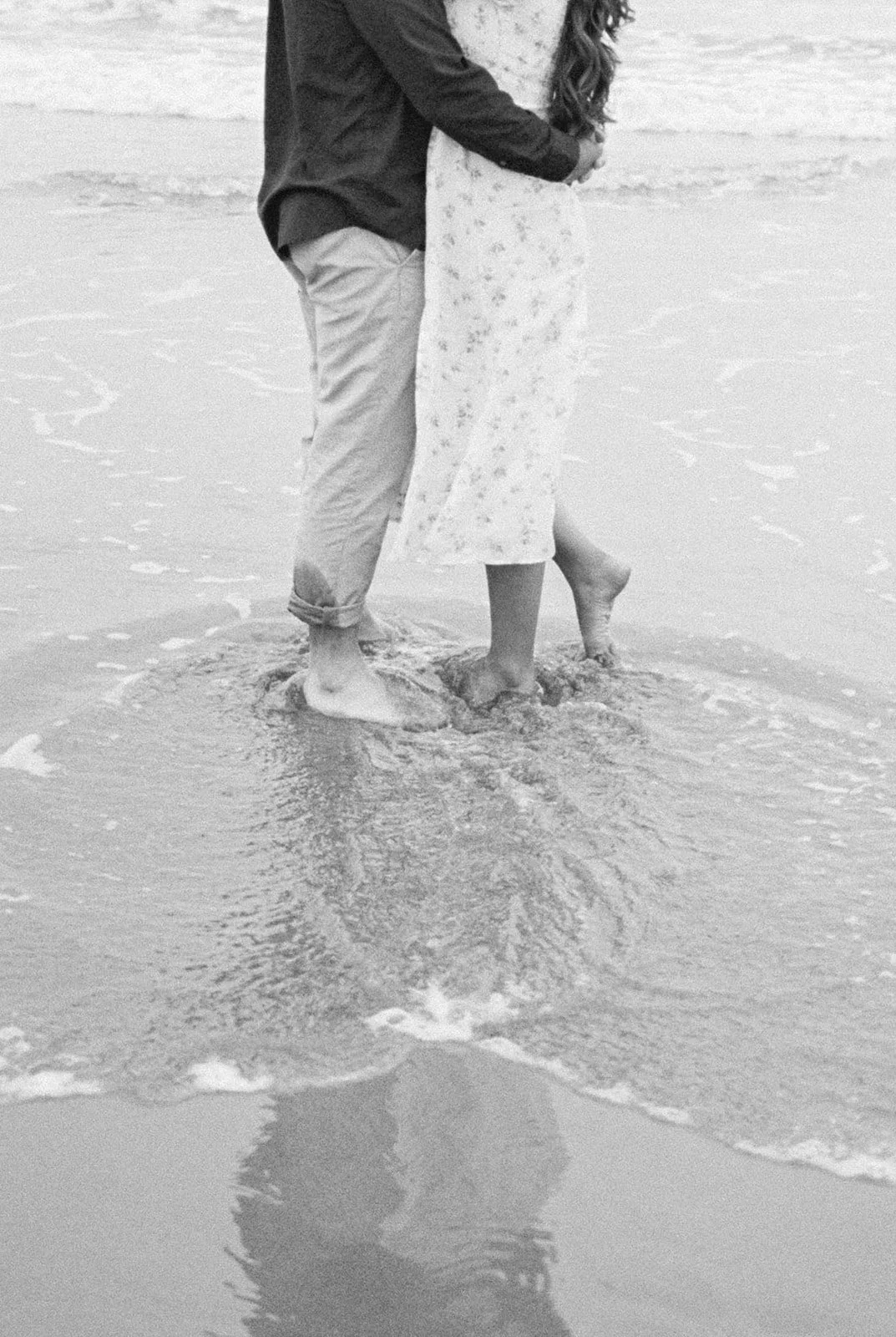 Close-up of couple’s feet in shallow water with ripples and reflection on beach