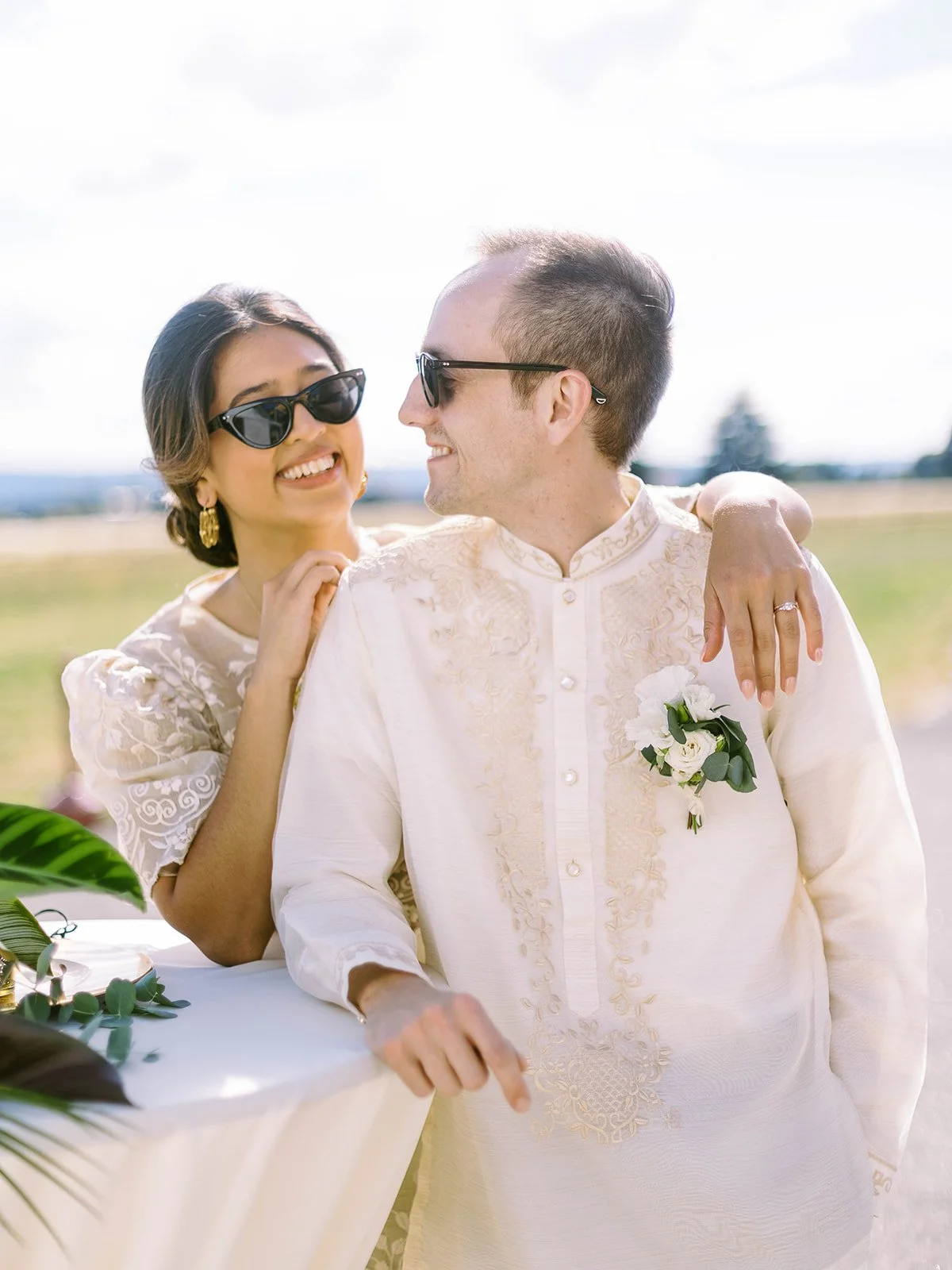 Bride and groom in sunglasses smiling at each other while leaning against a cocktail table at the airfield
