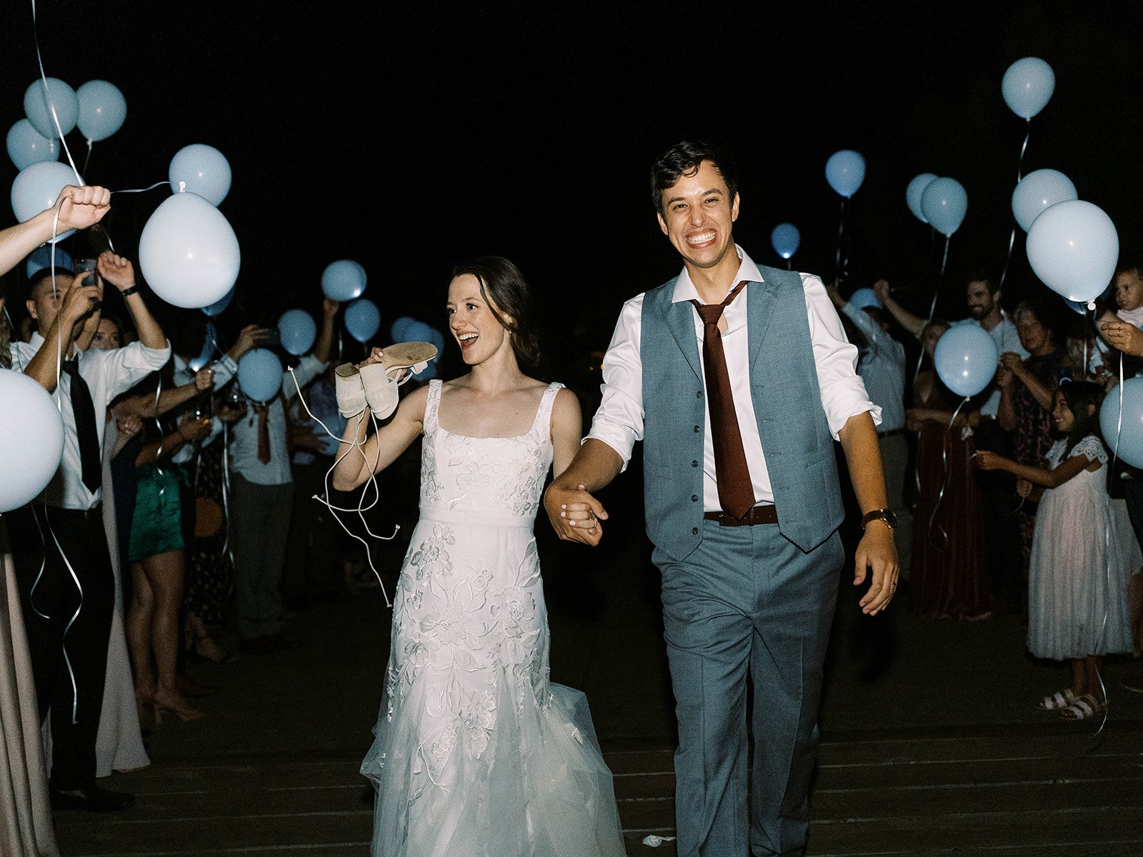 Bride and groom smiling as they walk through guests holding white balloons at the end of the wedding night.