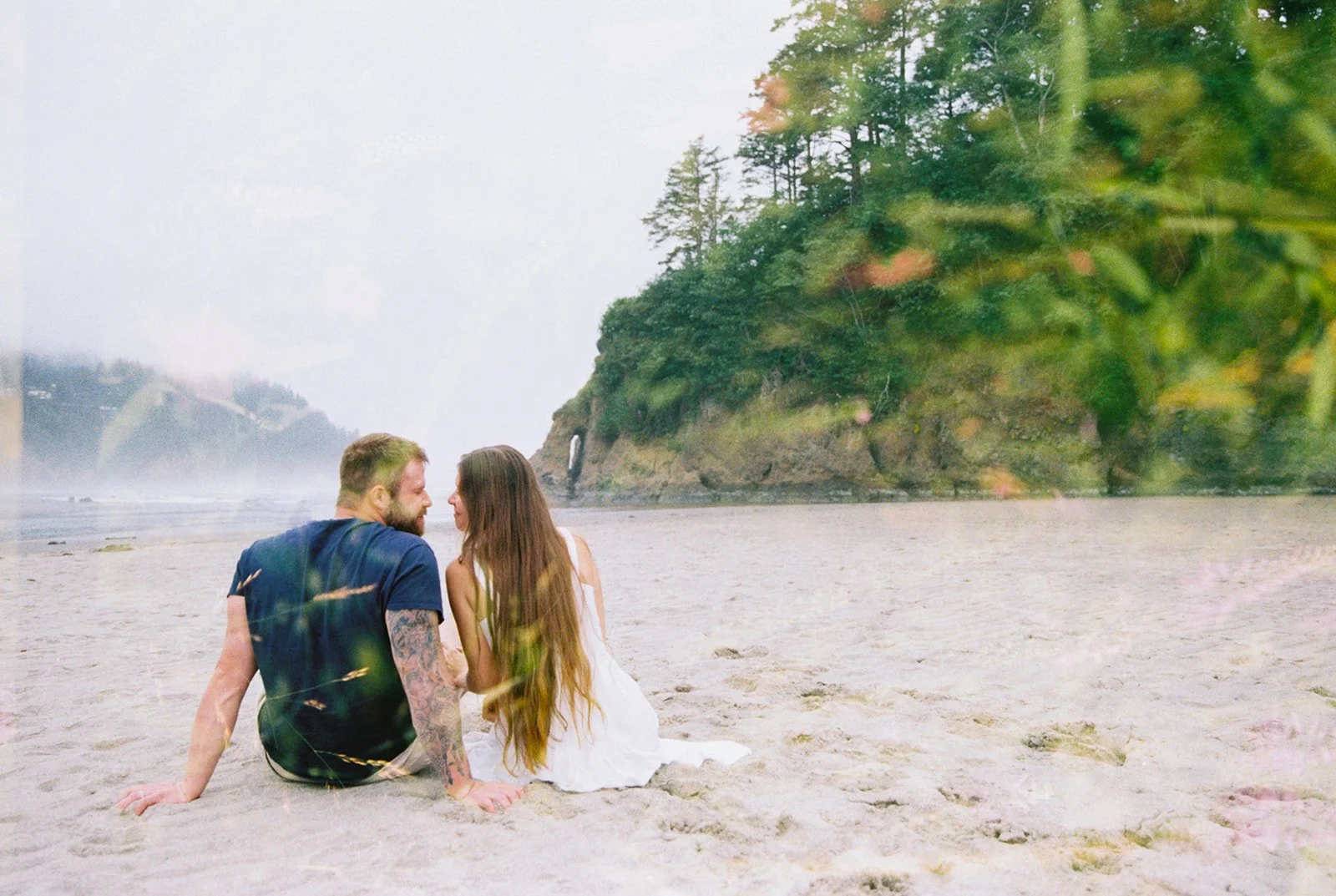 Couple sitting close together on beach with soft floral overlay and coastal cliffs in background.