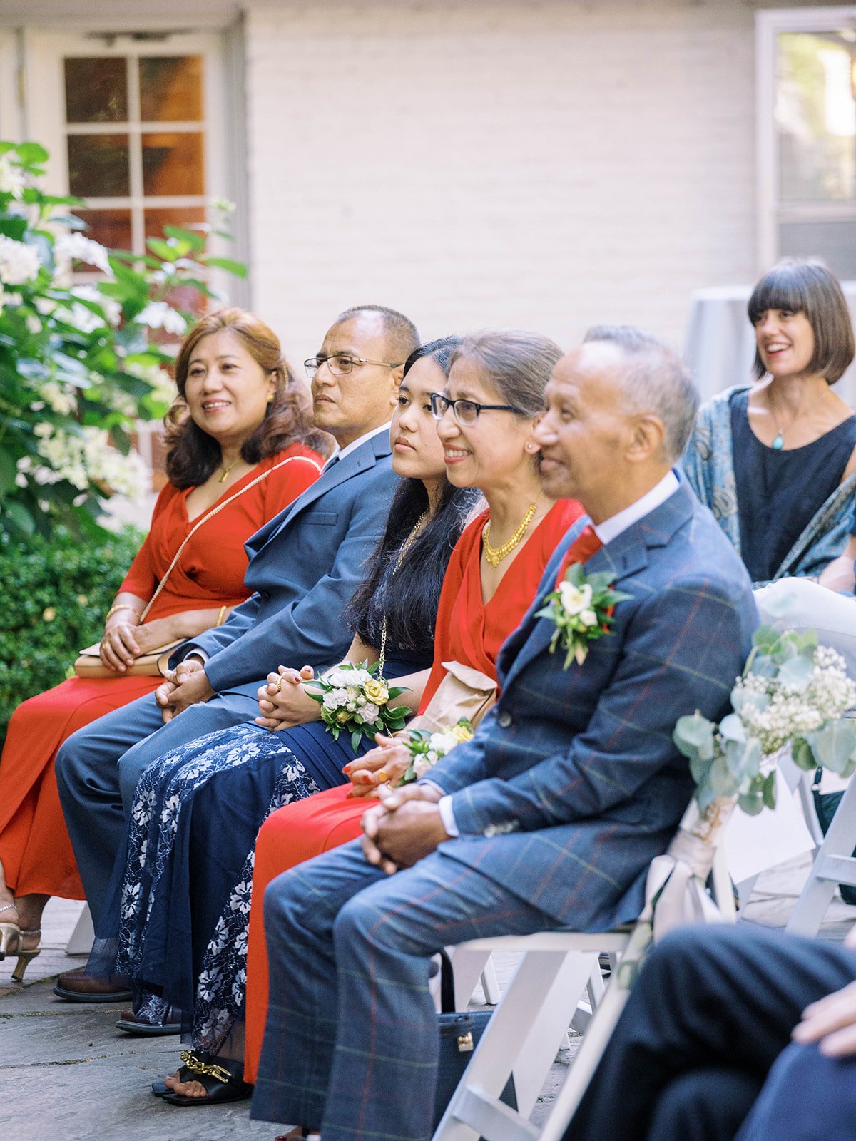 Wedding guests seated in the garden watch the ceremony from white folding chairs.