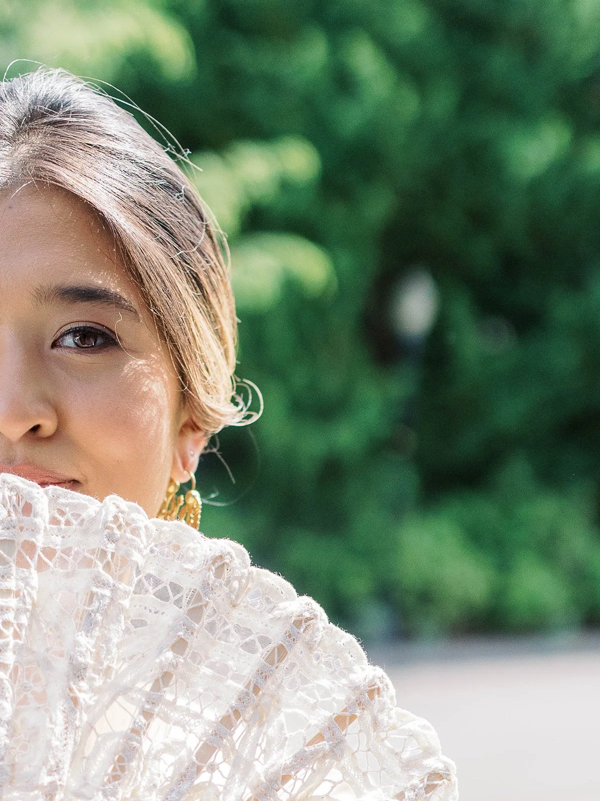 Close-up portrait of the bride peeking over a white lace fan, wearing gold earrings with green foliage behind her
