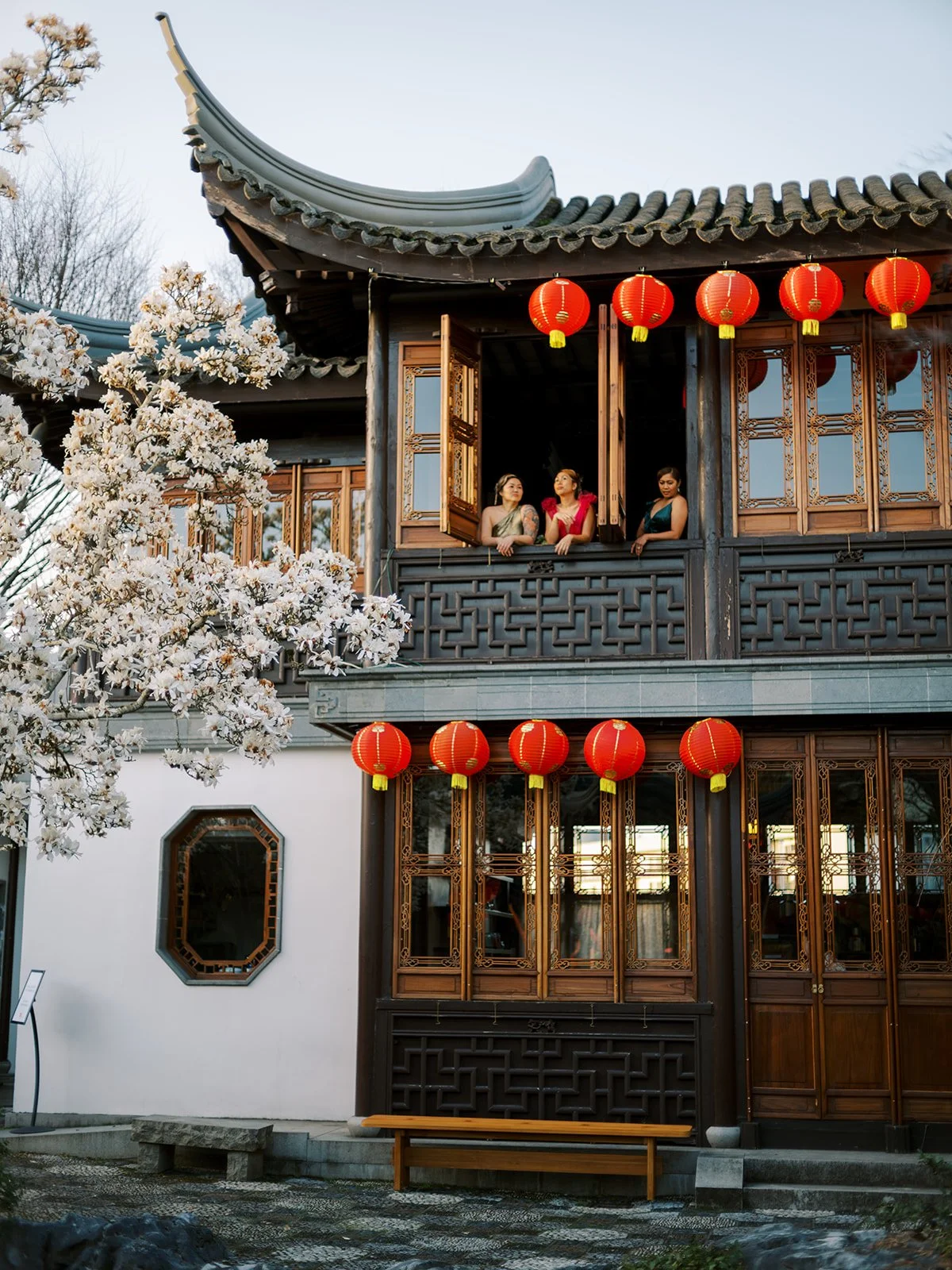 Three women in couture gowns standing on a balcony beneath red lanterns at Lan Su Chinese Garden in Portland during a bachelorette celebration.