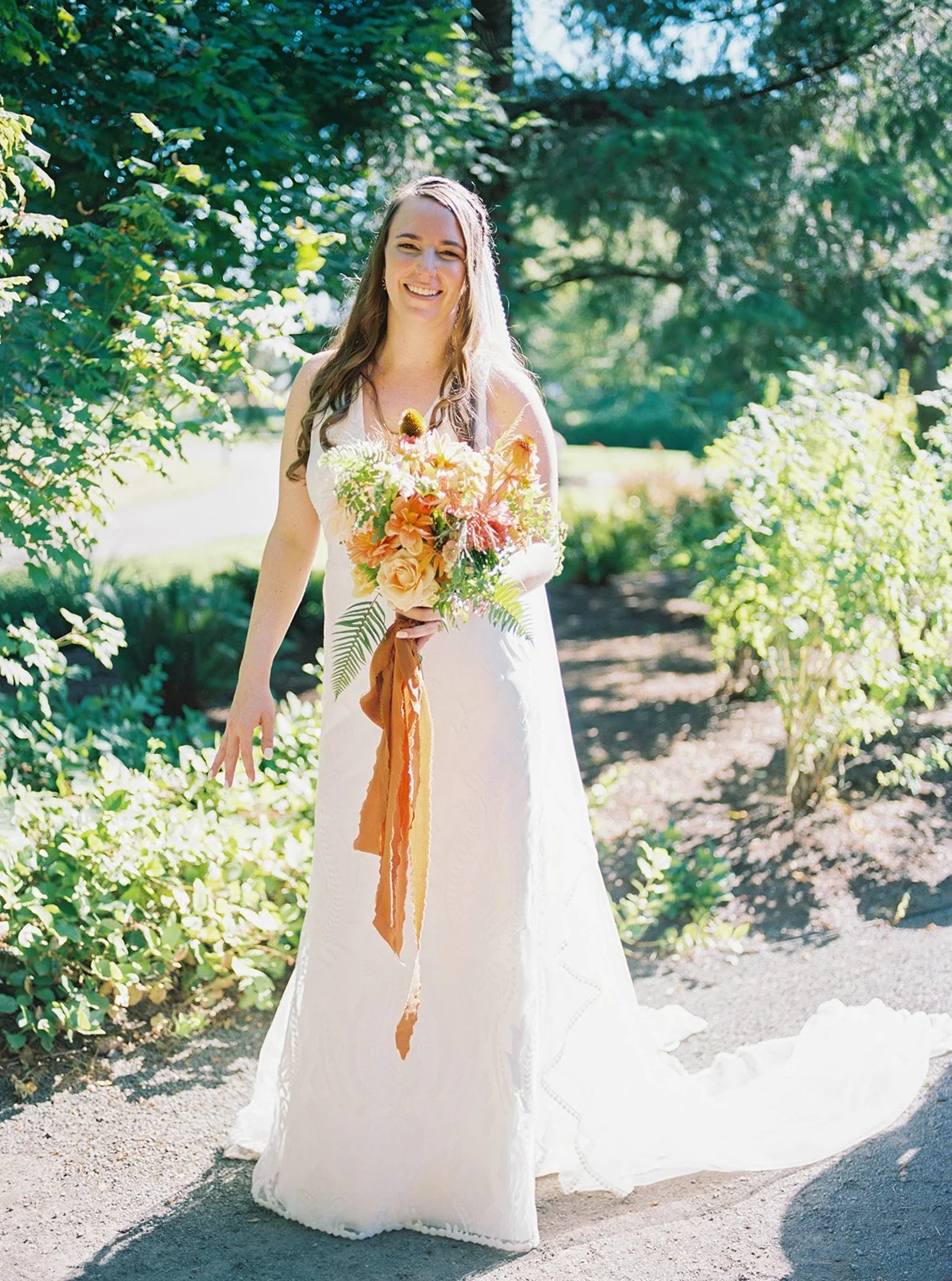 Bride in white gown holding colorful bouquet walking along sunlit garden path