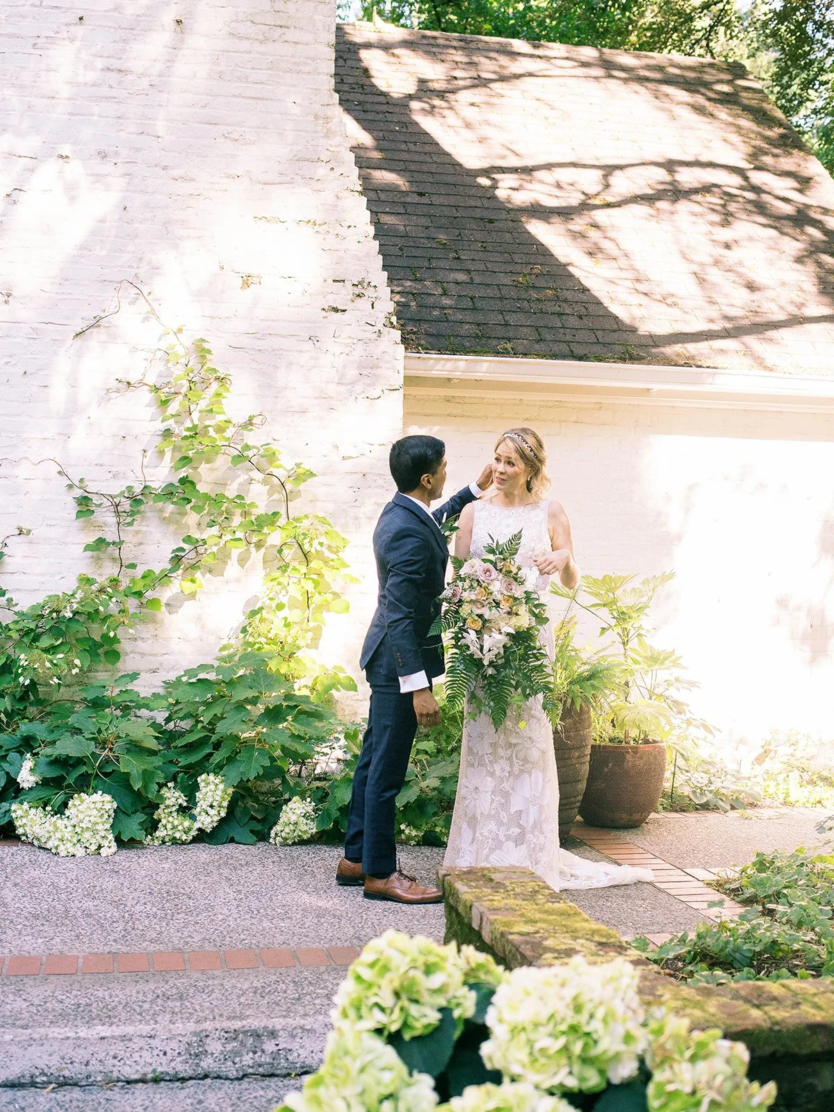 Groom gently touches the bride’s face during a quiet moment beside a garden wall.