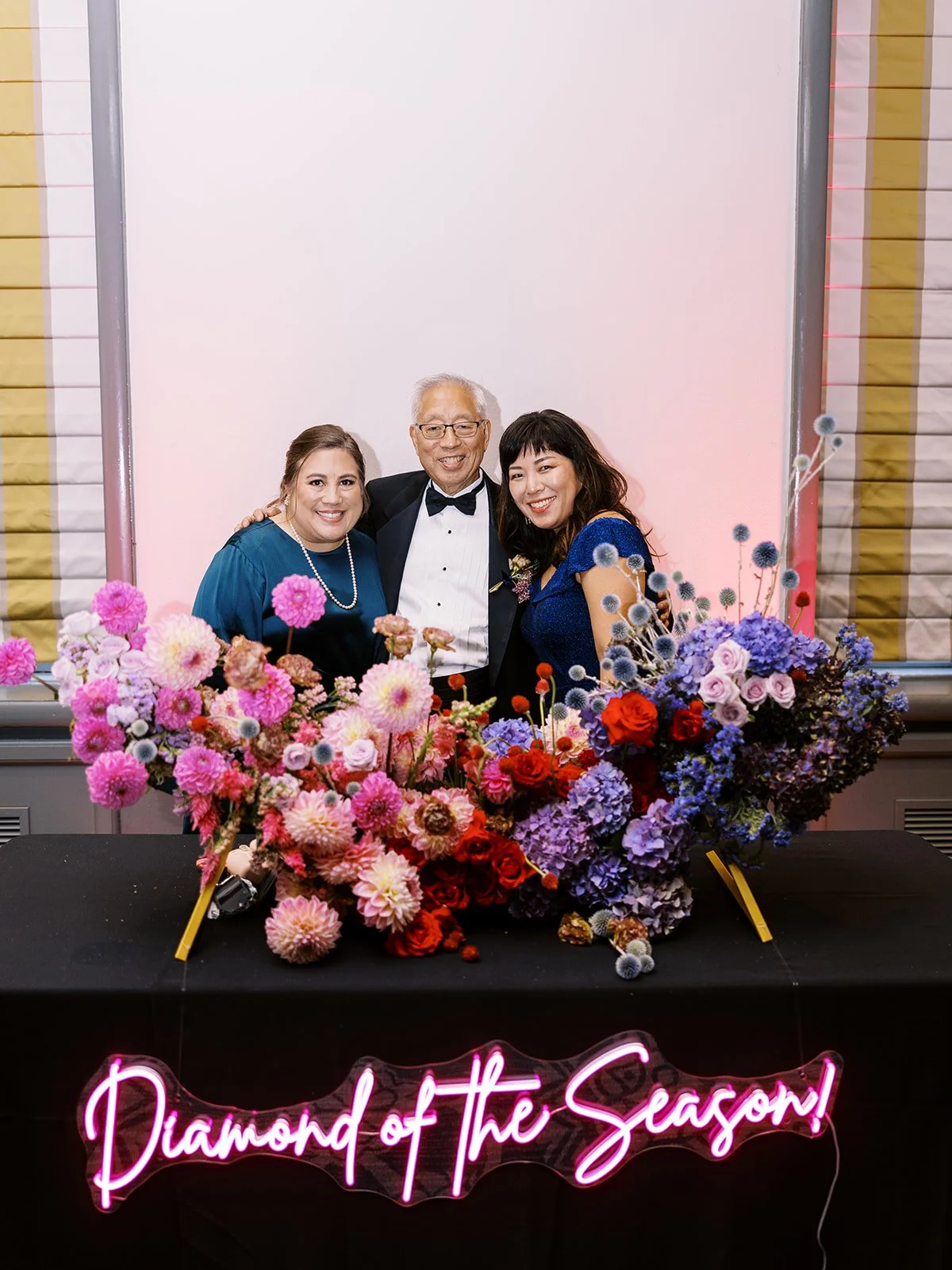 Three guests pose behind a vibrant floral installation with pink, red, and purple blooms and a neon “Diamond of the Season” sign.