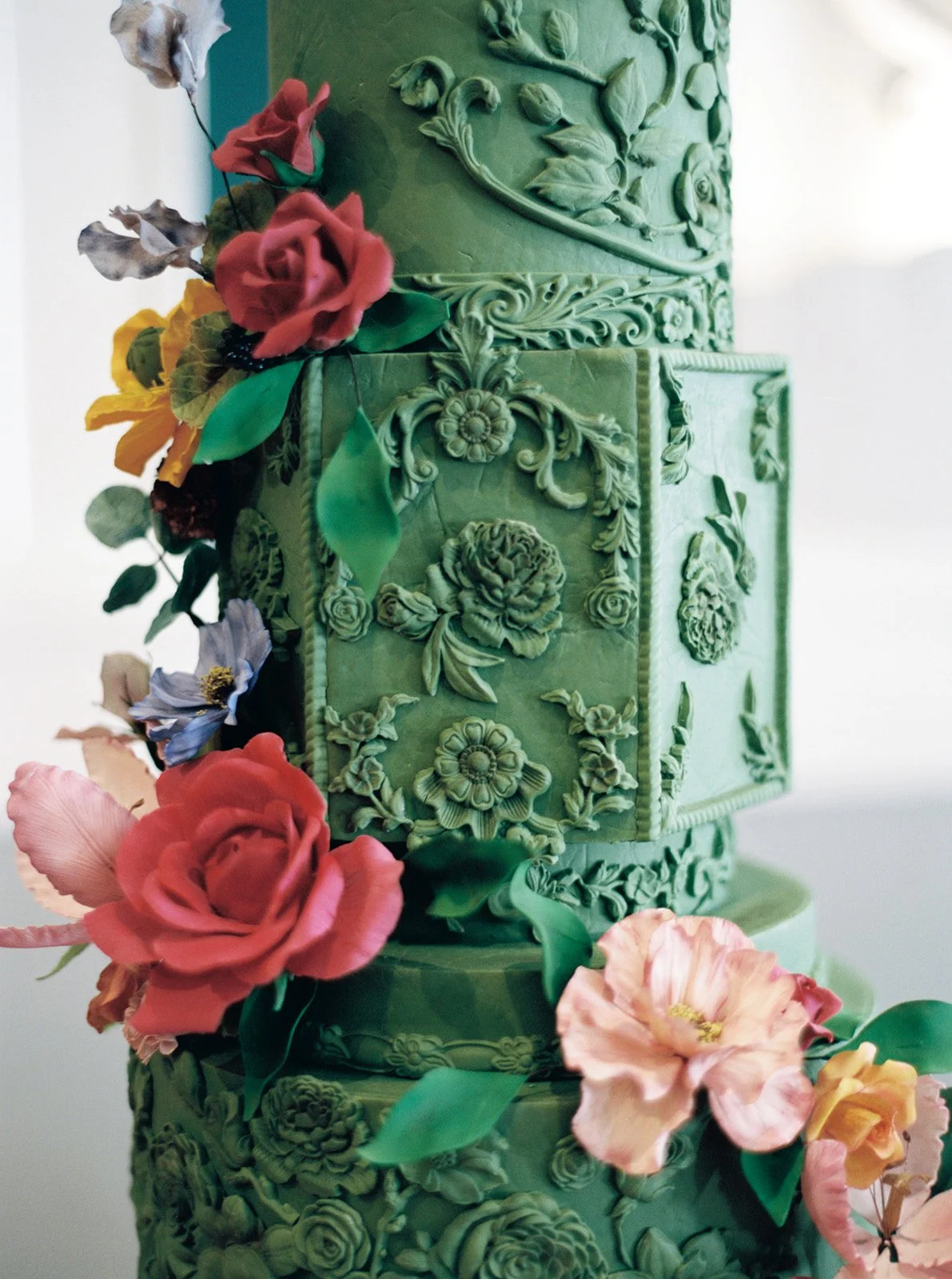 Details of embossed floral patterns on a green tiered cake accented with red and pastel sugar flowers