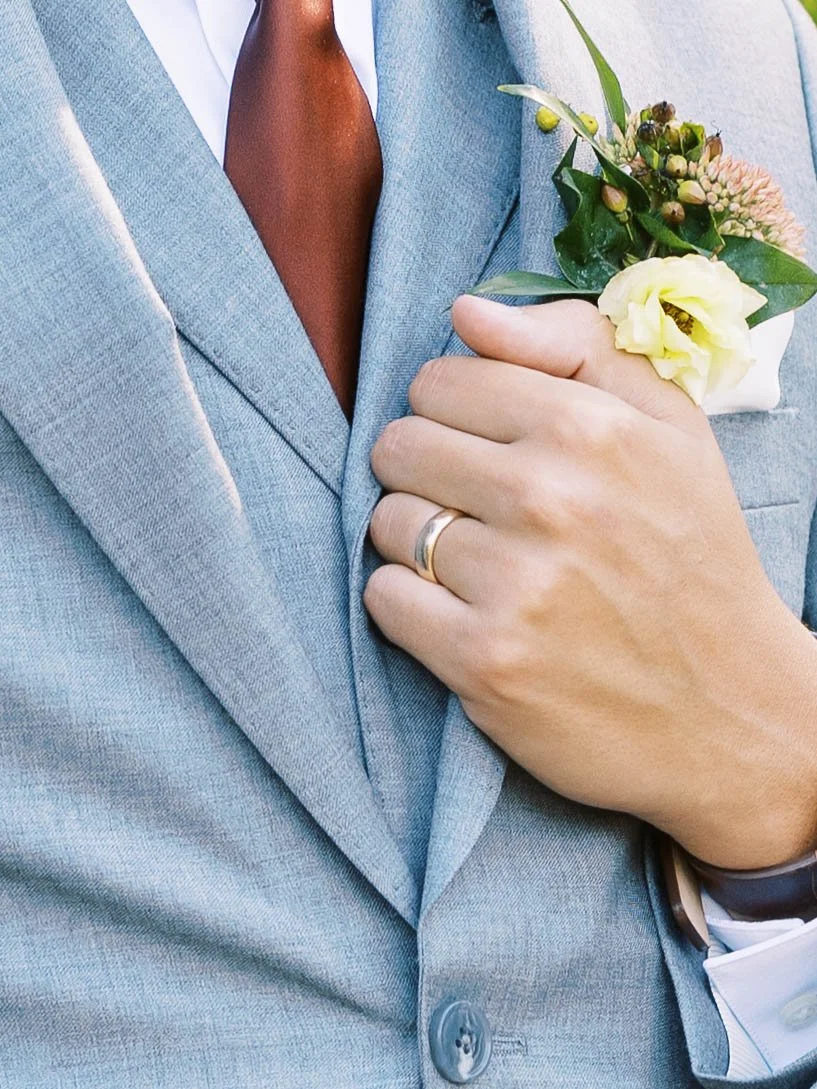 Close-up of a gray wedding suit with a rust-colored tie, floral boutonniere and wedding band.