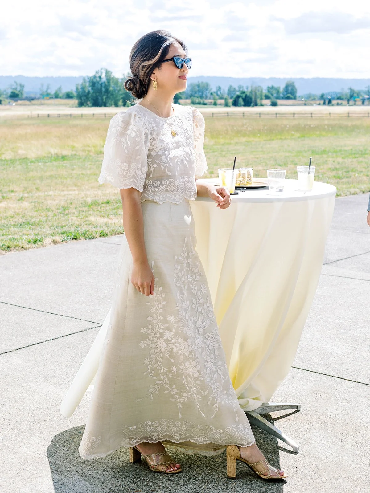 Bride in embroidered Filipino gown and cat-eye sunglasses standing at a cocktail table overlooking the Pearson Airfield runway
