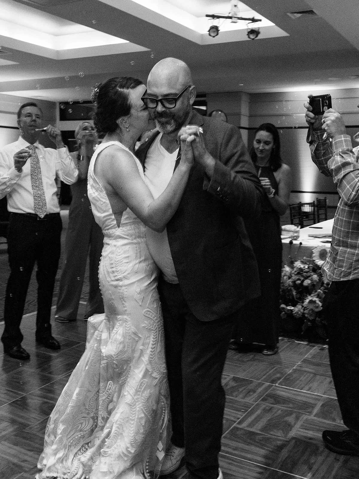 Black and white photo of the bride kissing the groom's cheek as they dance, guests watching and capturing the moment with phones and cameras