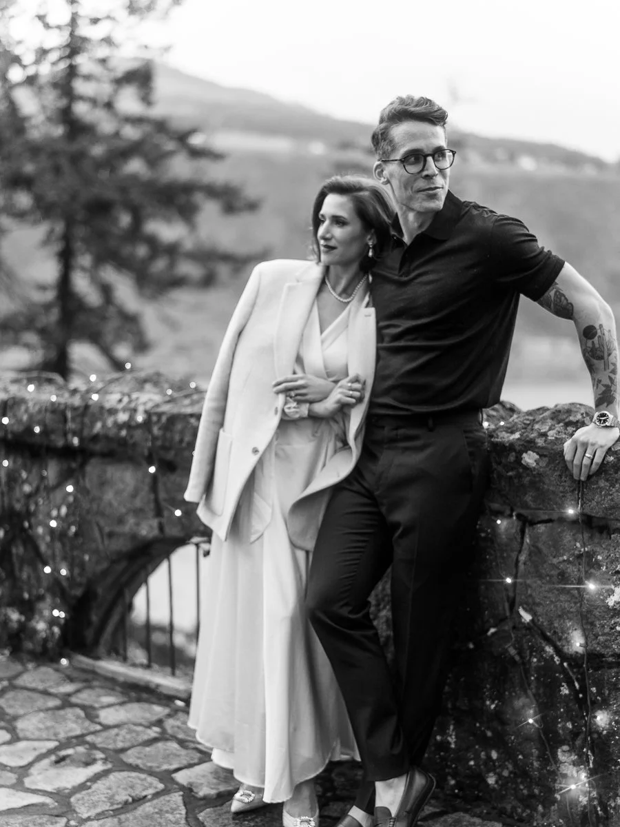 Black and white portrait of a couple posed against a moss-covered stone wall with a mountain river view and fairy lights.