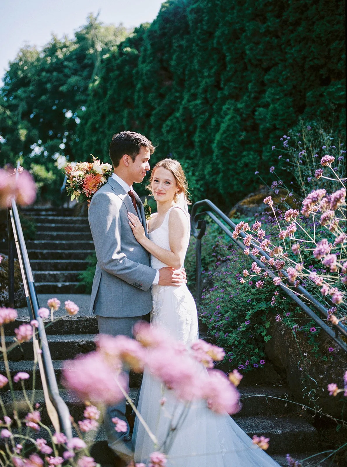 Bride and groom standing close together on garden steps, framed by soft pink flowers and greenery.