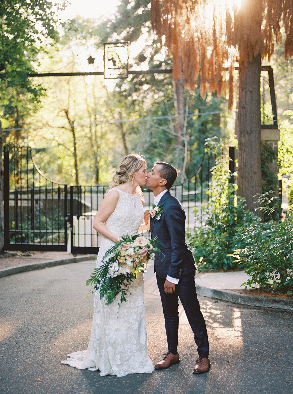 Bride and groom kiss near the garden entrance with sunlight filtering through the trees.