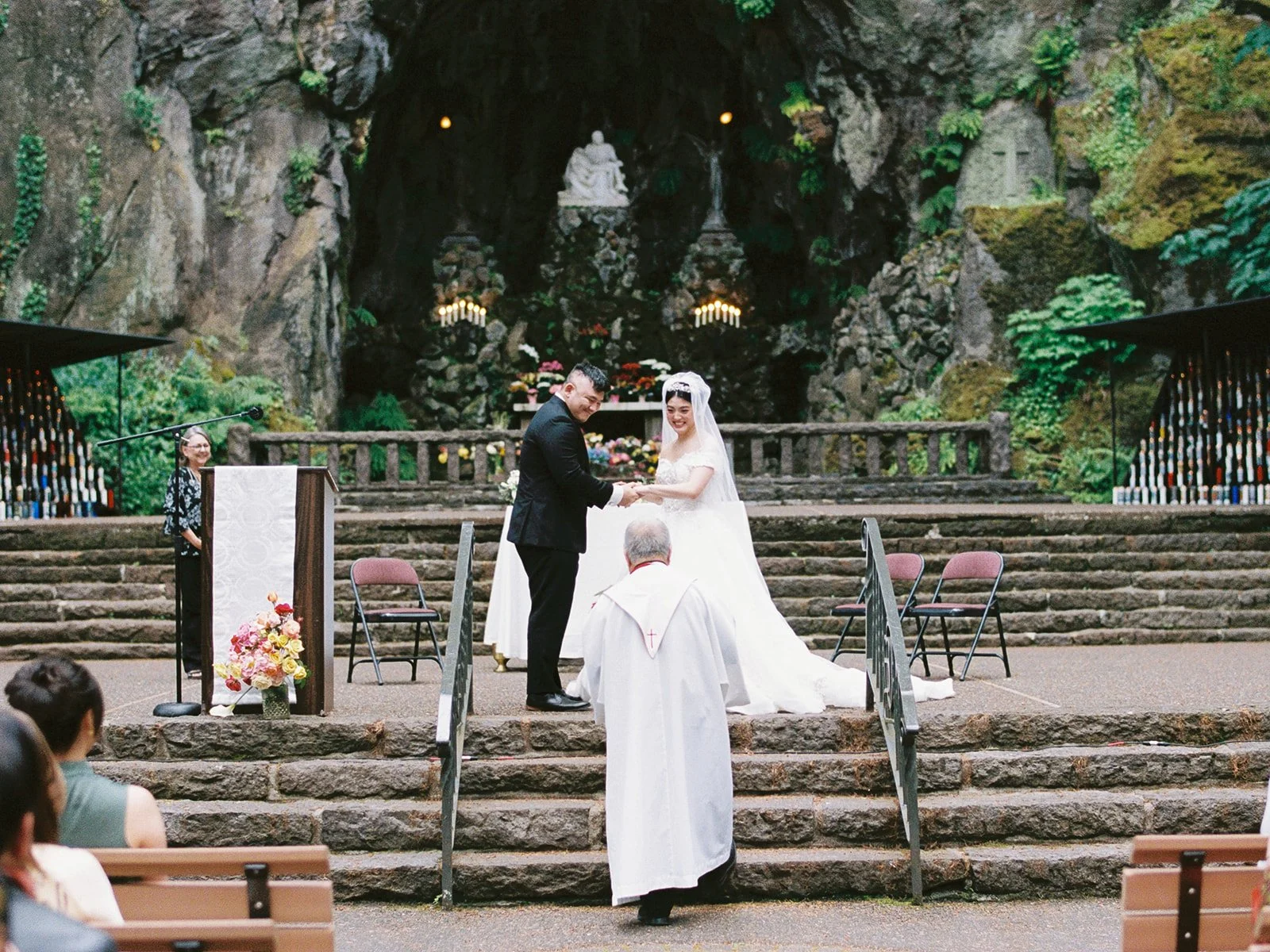 The groom places a ring on the bride's finger at The Grotto outdoor altar, a priest in white vestments officiating, the cave shrine and colorful flowers behind them