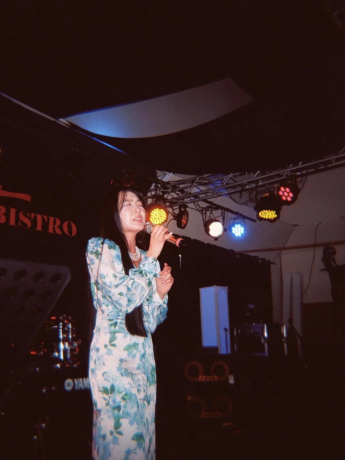 A woman in a blue floral áo dài singing with eyes closed on the Mekong Bistro stage under warm stage lighting