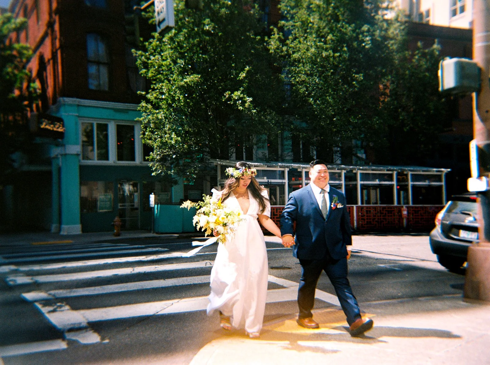 Bride in a white dress and colorful flower crown holds hands with her partner while crossing a sunny Portland street.