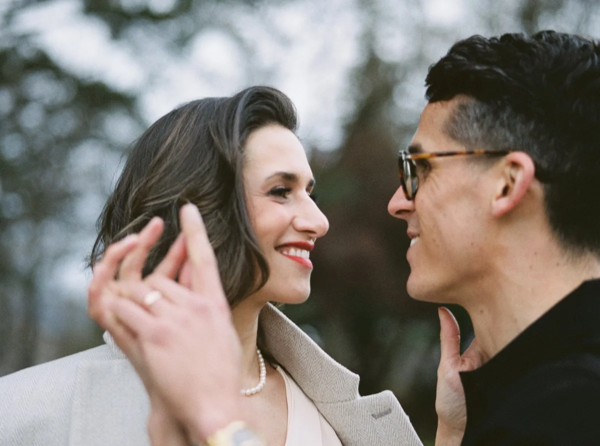 Close-up of couple smiling at each other, woman in pearl necklace and cream coat, man in glasses and black shirt