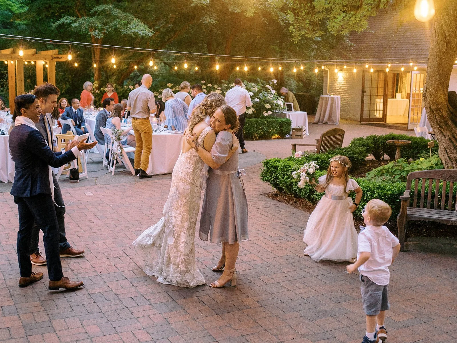 Bride hugs a wedding guest as children gather nearby beneath warm evening string lights.
