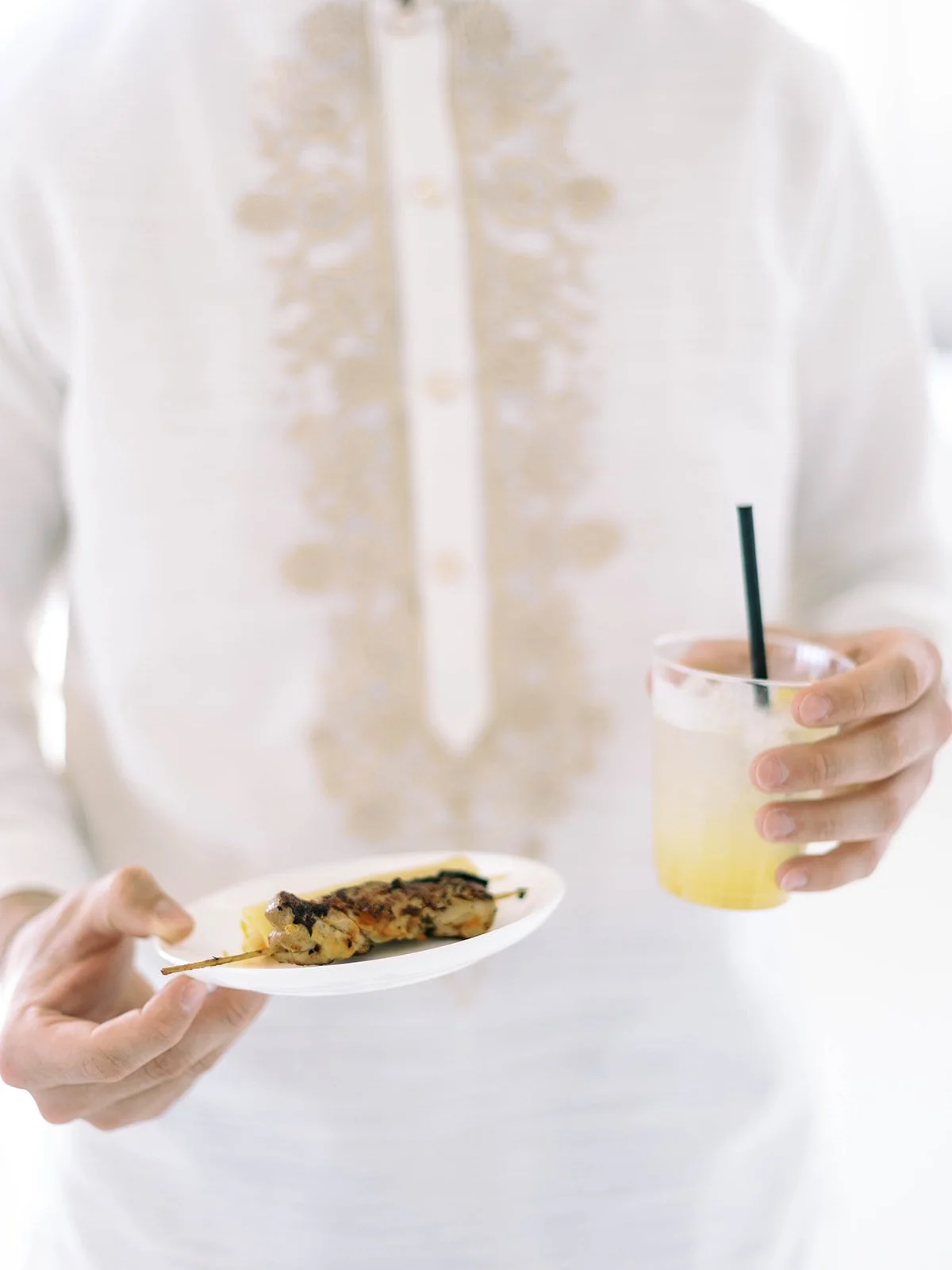Close-up of a wedding guest in barong tagalog holding a plate of chicken skewers and a cocktail drink

