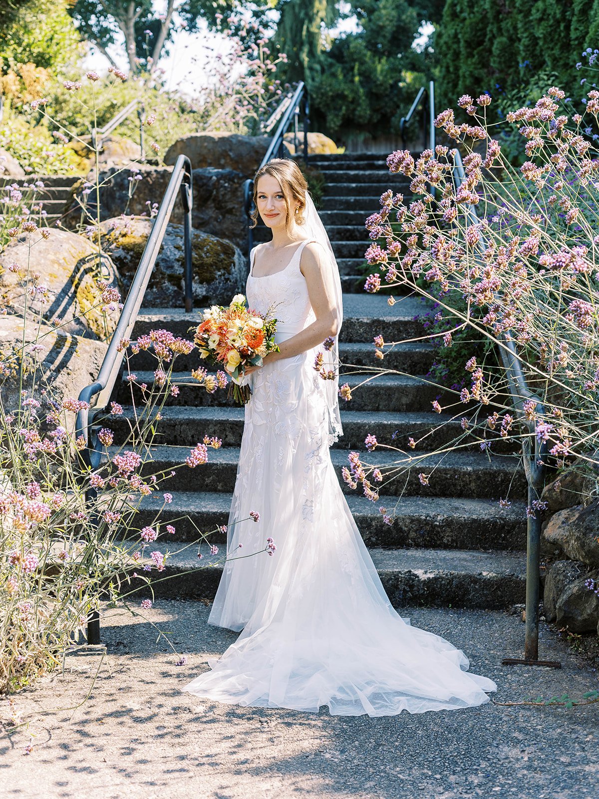Bride holding a warm-toned bouquet while standing on stone garden steps surrounded by purple flowers.