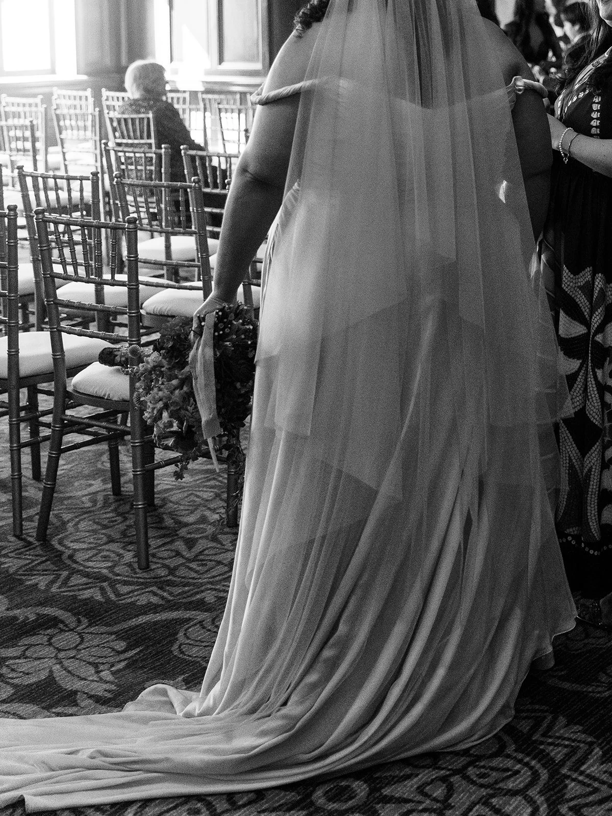 Bride walking down aisle holding bouquet, gown and veil flowing behind in indoor venue.