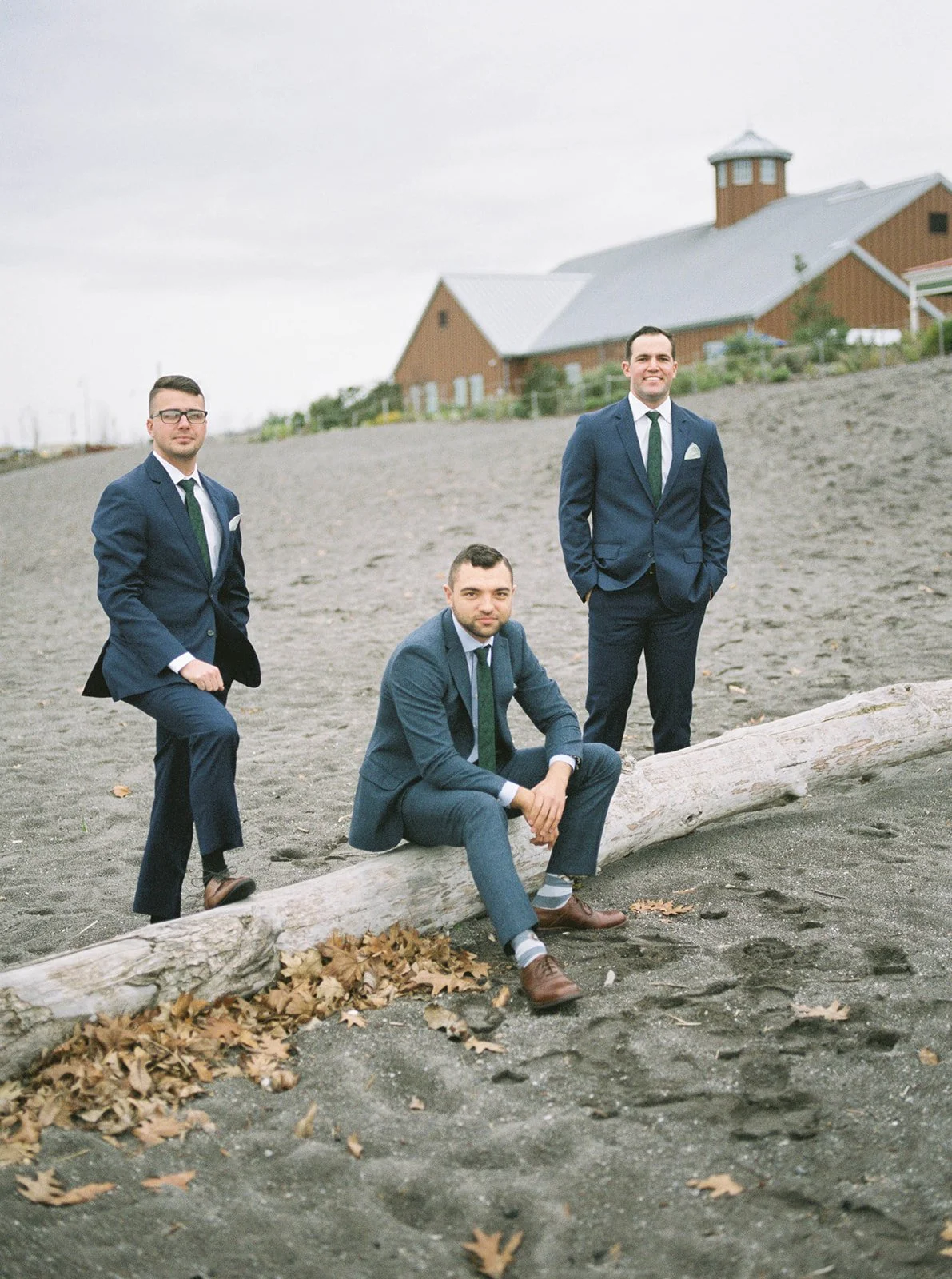 Groom and two groomsmen pose beside driftwood on the sandy riverfront in matching blue suits.