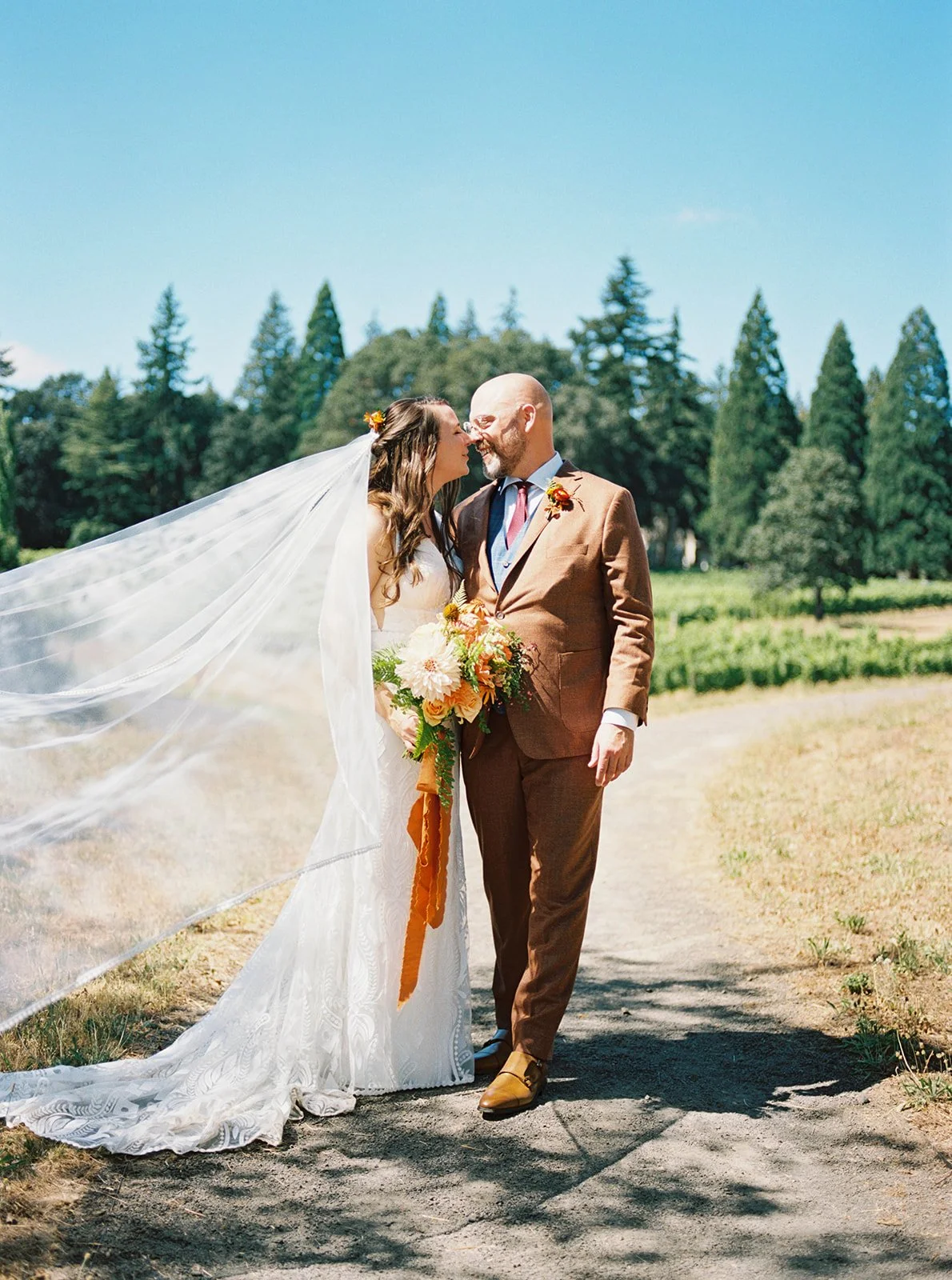 Bride and groom walking together on path, sharing a kiss with flowing veil