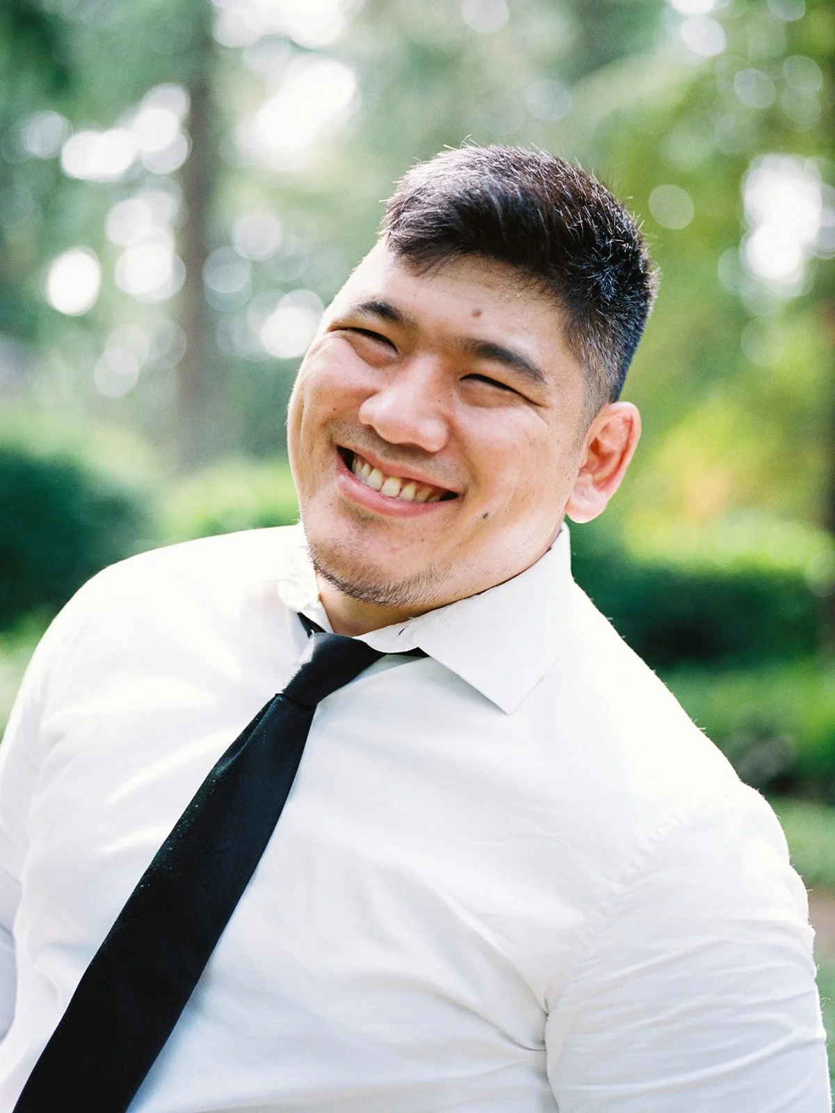 Close-up portrait of the groom laughing broadly in dappled forest light at The Grotto, white shirt and black tie