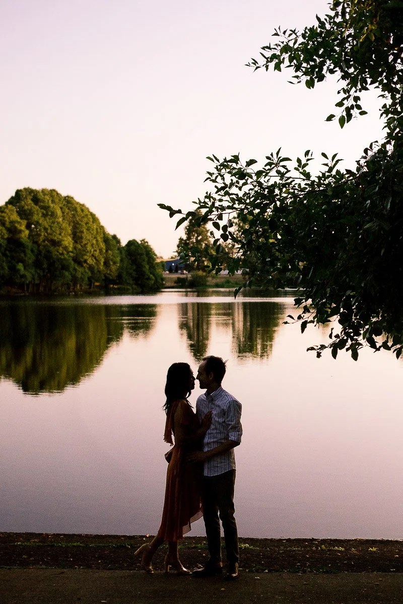 Silhouette of couple standing by calm lake at sunset, framed by trees and warm evening light reflections
