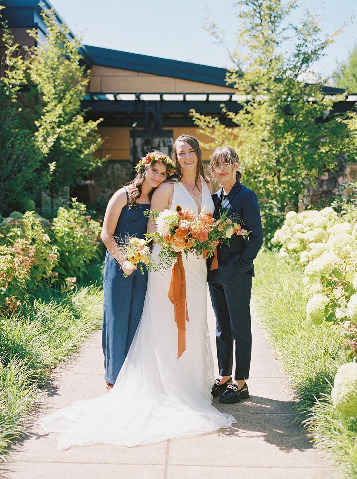 Bride standing with two wedding attendants in a garden, holding warm-toned floral bouquets.