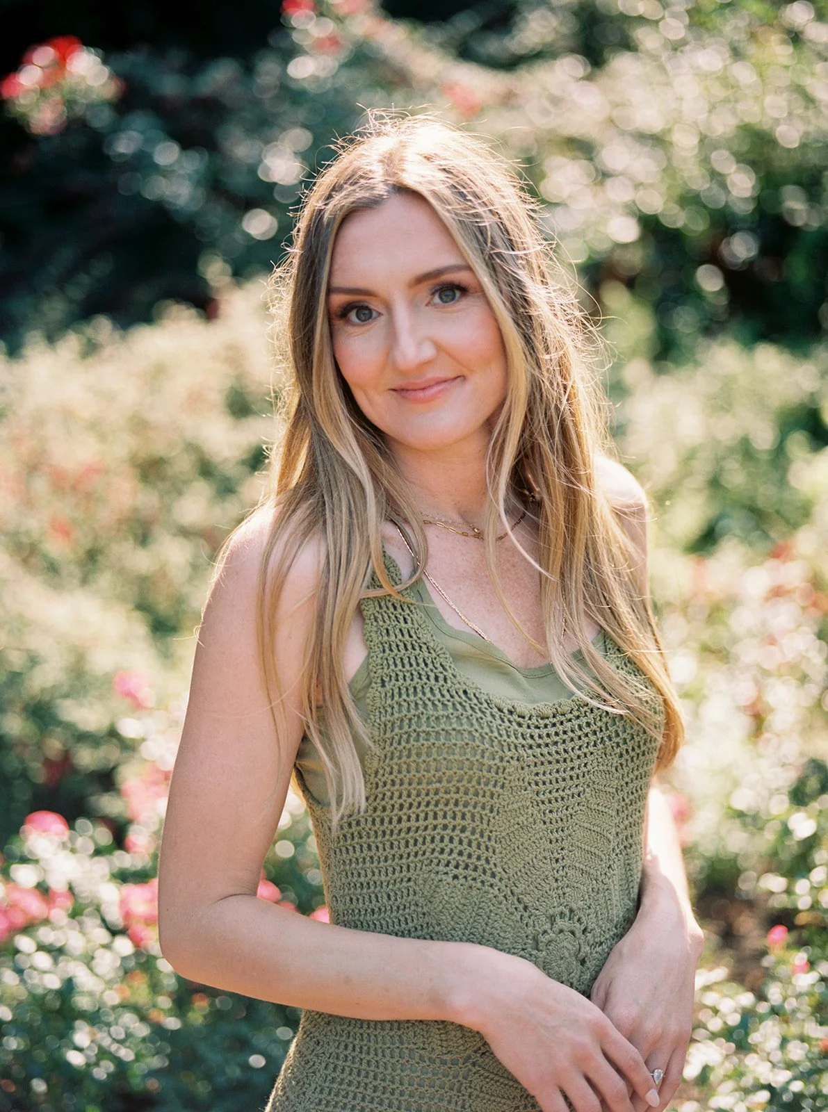 A solo portrait of a woman in a green crochet dress smiling softly, surrounded by blurred pink roses in warm sunlight.