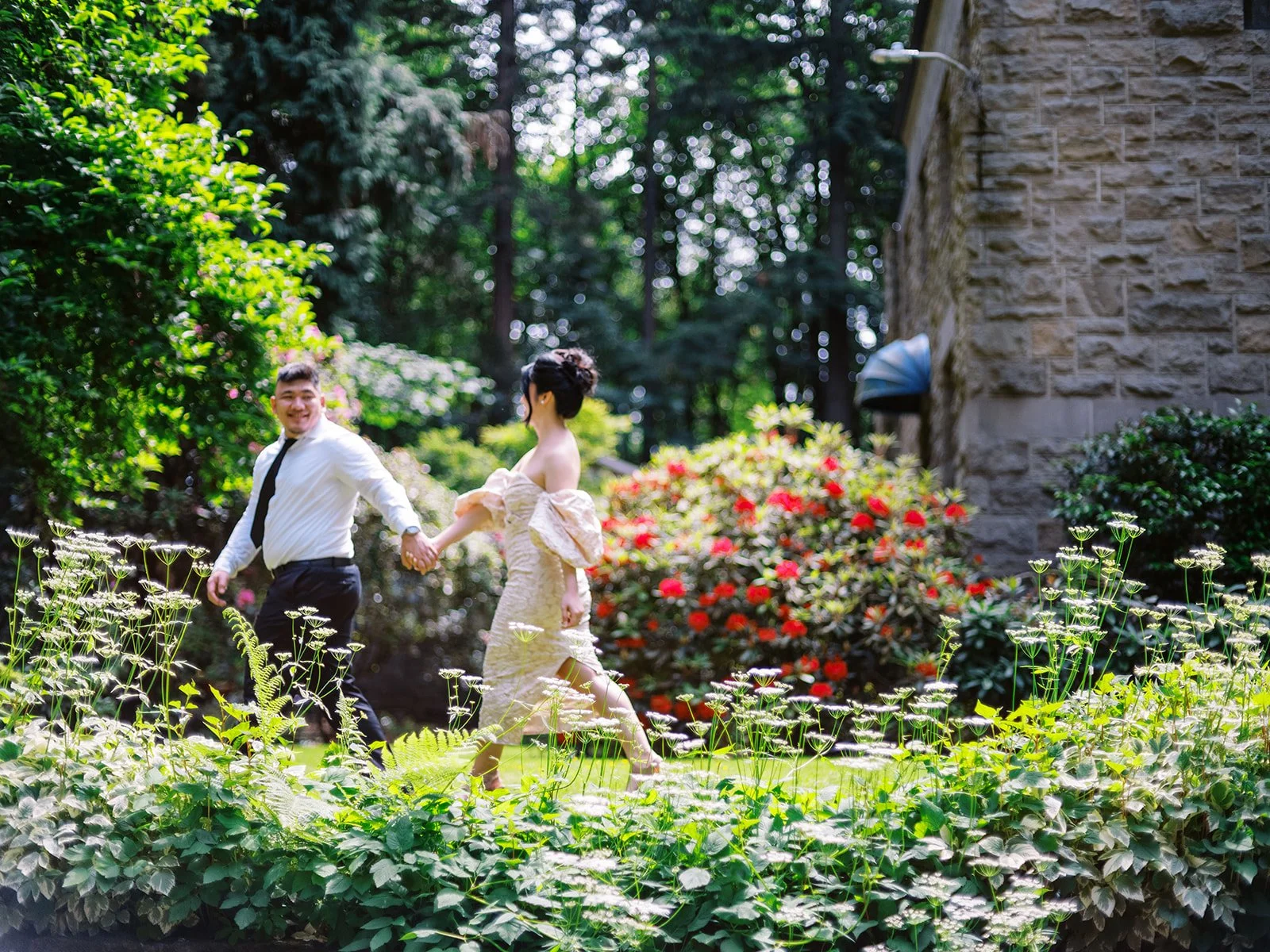 Bride and groom running hand-in-hand through a sunlit garden at The Grotto, red rhododendrons blooming beside the stone chapel wall
