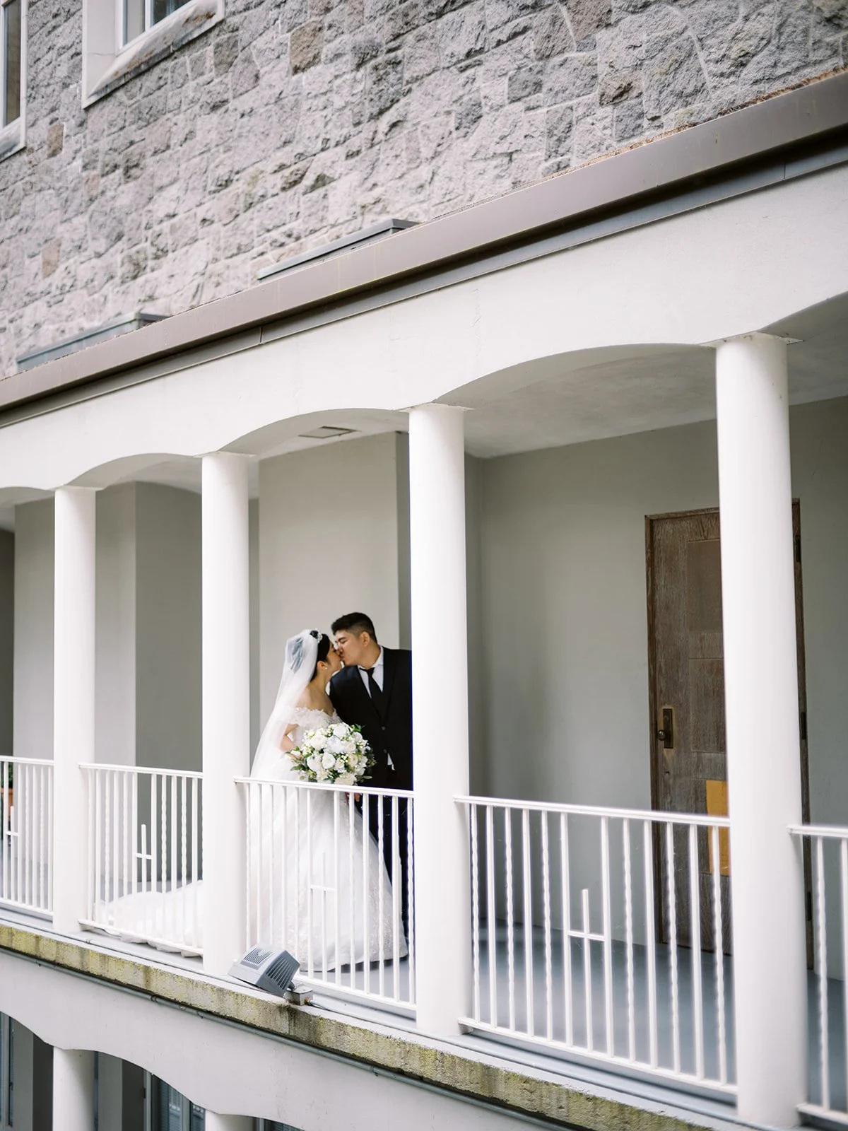 Wide shot of the bride and groom sharing a kiss on the colonnaded upper terrace of The Grotto's stone building, her white bouquet and ball gown train visible below the railing