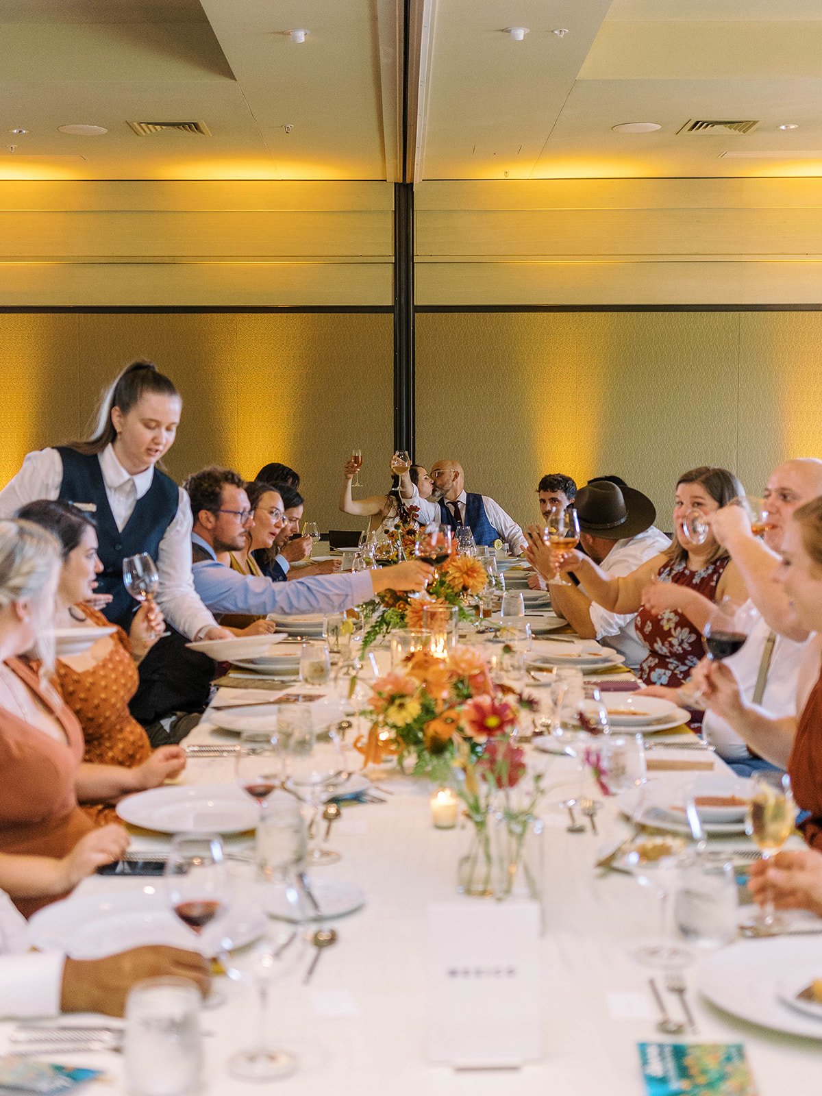 Guests at a long reception table raising glasses in a toast while the couple kisses at the head of the table, with a server attending nearby