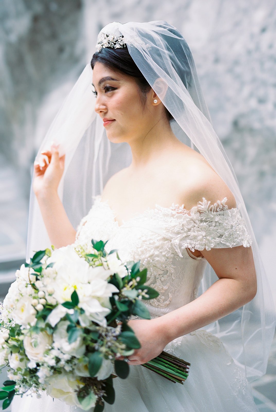 Profile portrait of the bride in an off-shoulder lace ball gown and tiara holding a white and green bouquet, The Grotto's stone wall behind her