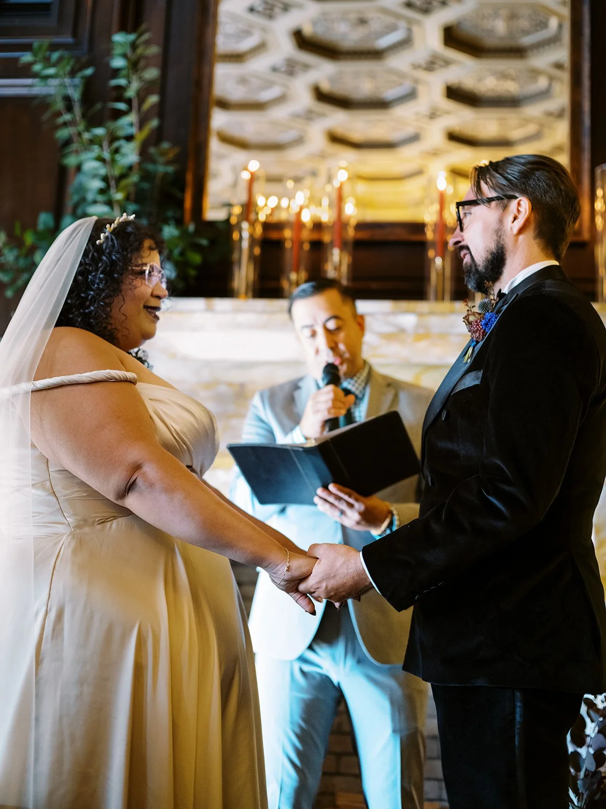 Close-up of couple holding hands during wedding ceremony as officiant reads vows in elegant indoor venue.
